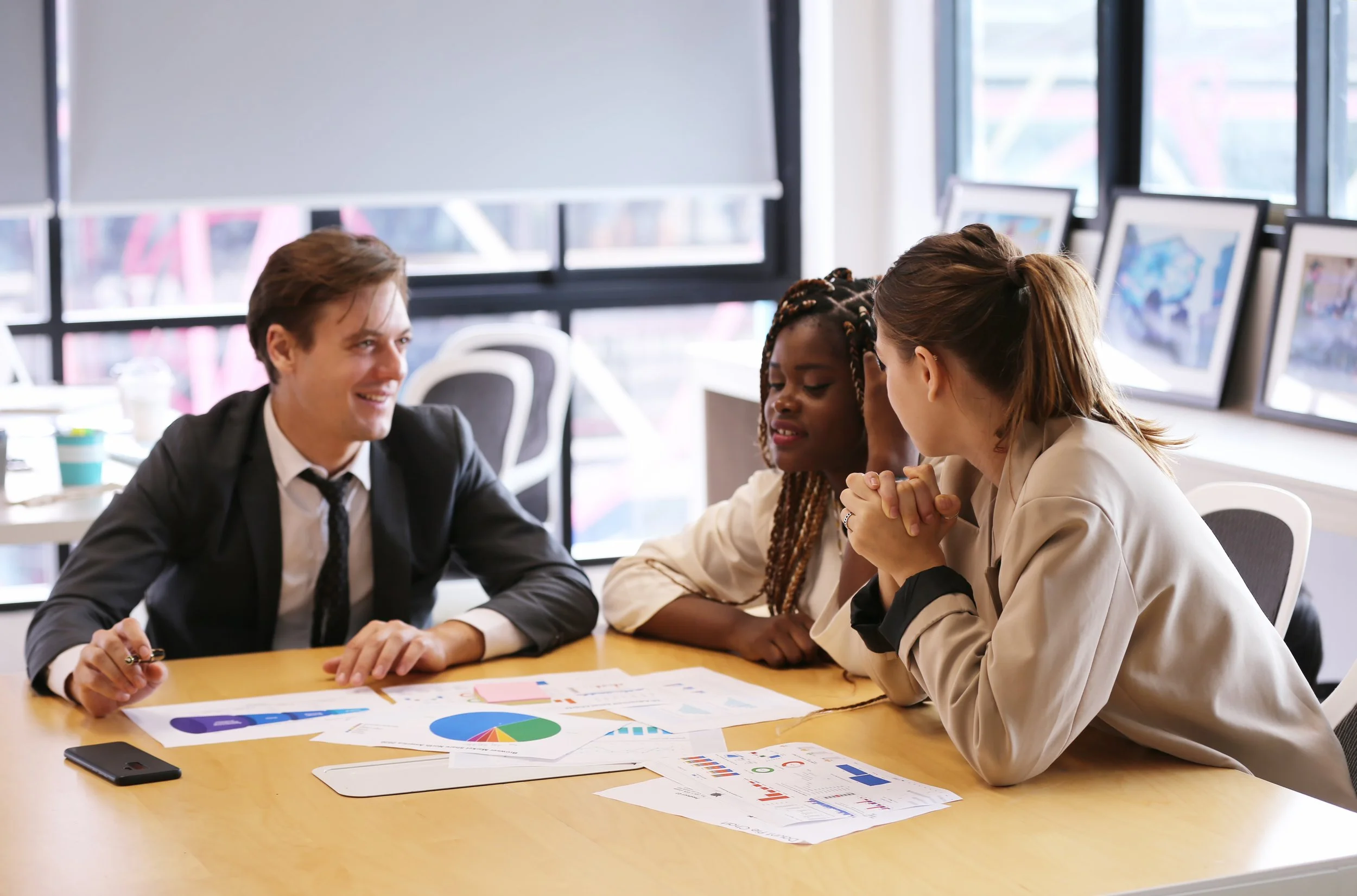 Three colleagues having a business meeting in a modern office, discussing documents with charts and graphs on a table.