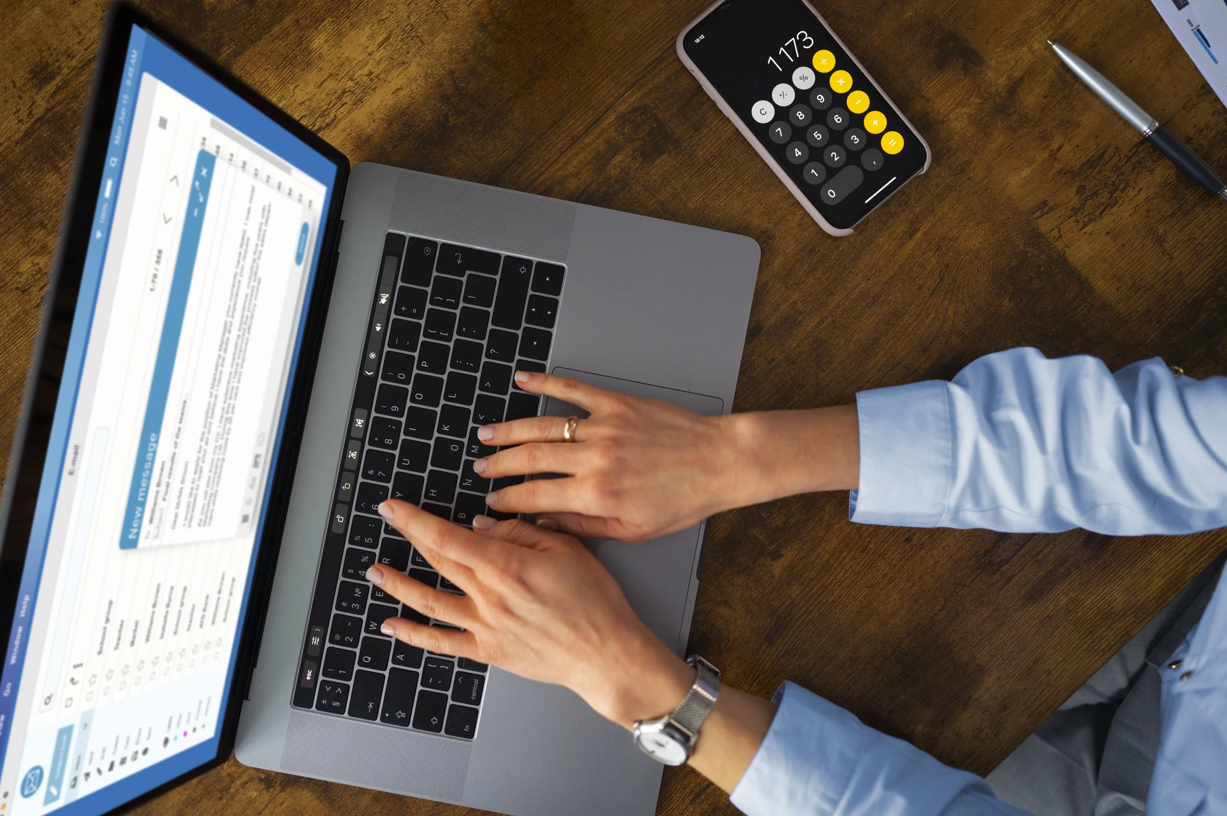 Person typing on a laptop keyboard, with a smartphone displaying a calculator app and a notepad with a pen on a wooden desk.