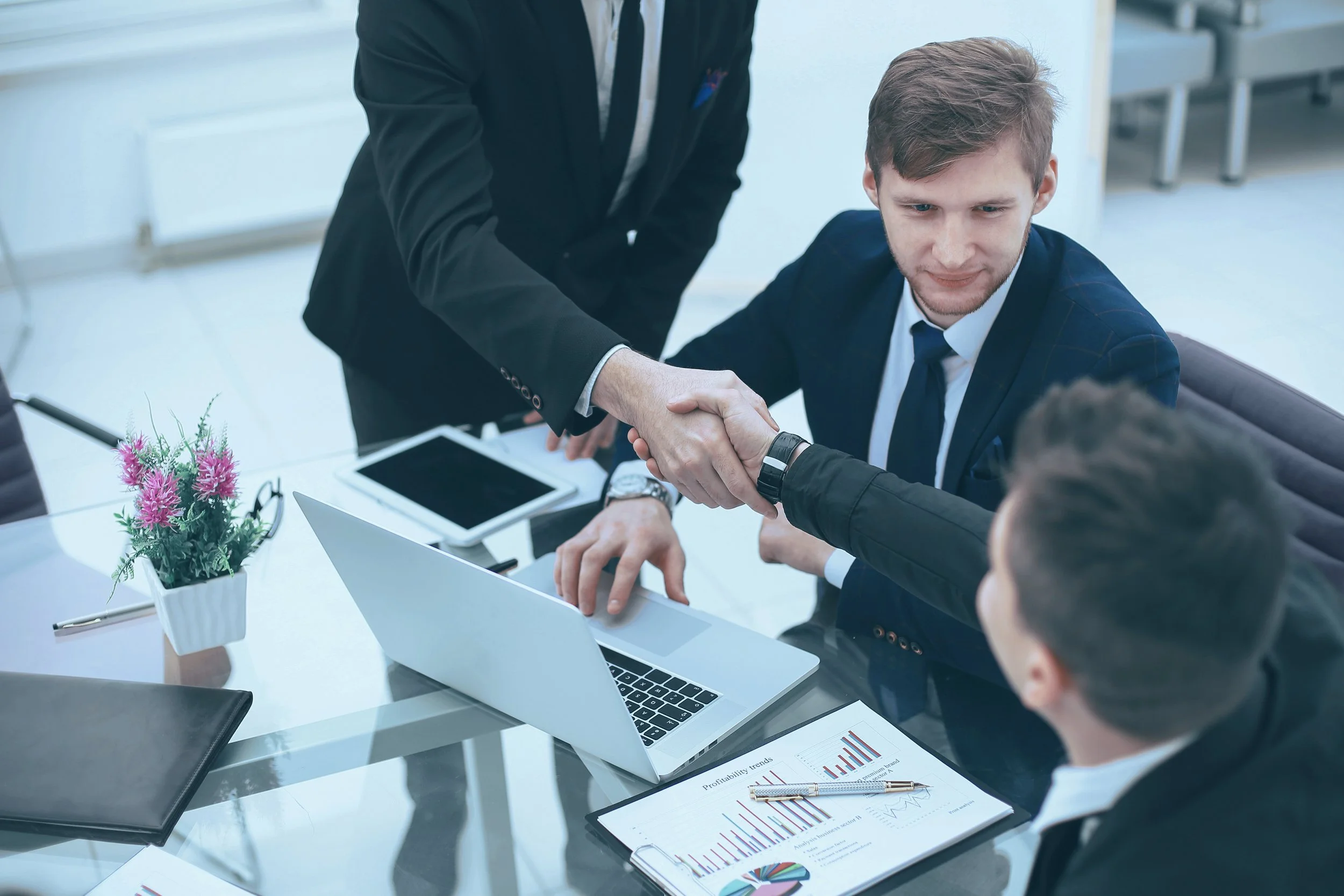 People shaking hands at a business meeting in a modern office with laptops and charts.