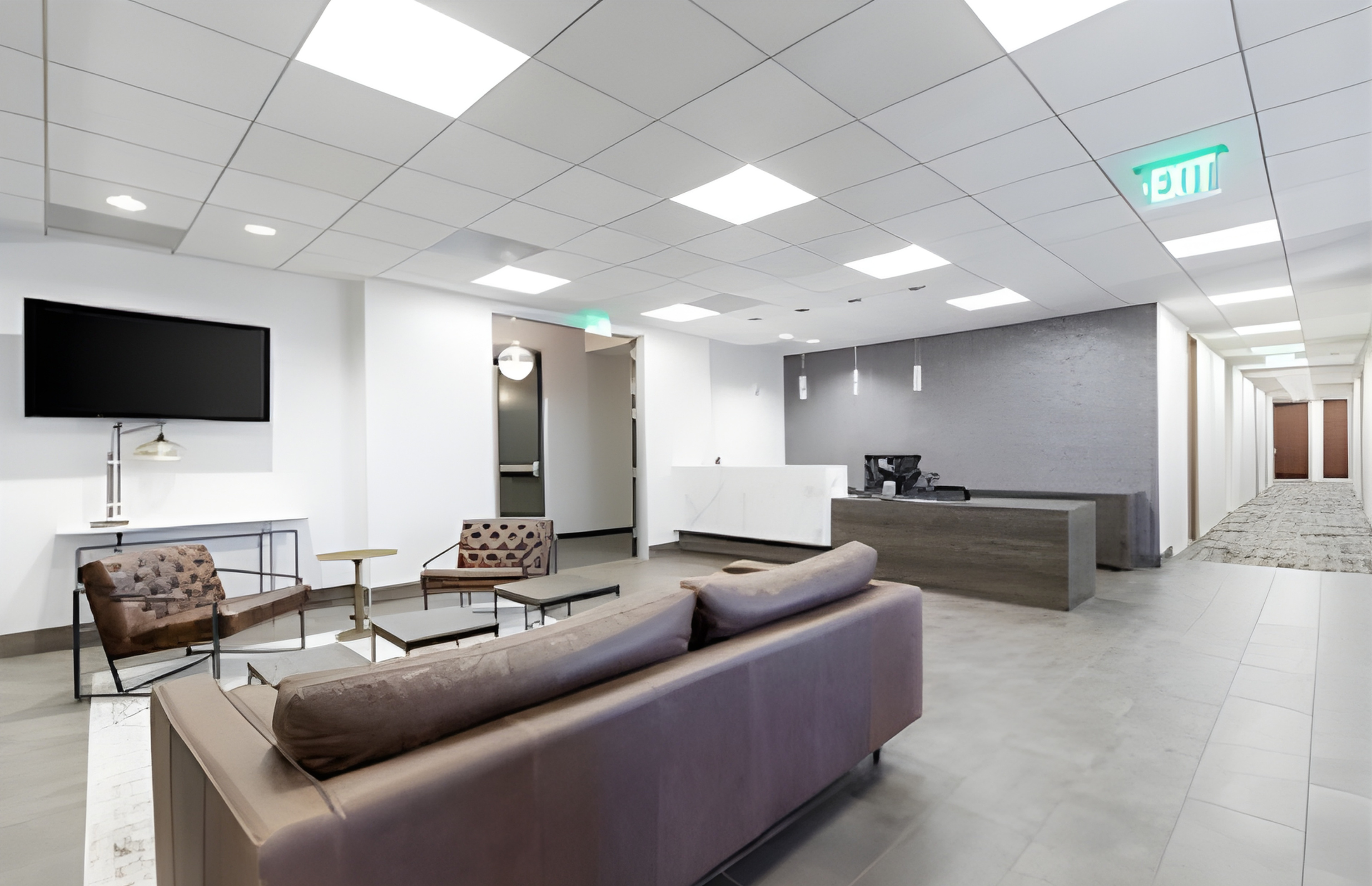 Modern hotel lobby with white walls, tiled flooring, and a gray accent wall. Contains a purple sofa, two patterned armchairs, a glass coffee table, a large flat-screen TV, and a reception desk. A hallway with closed doors extends to the right.