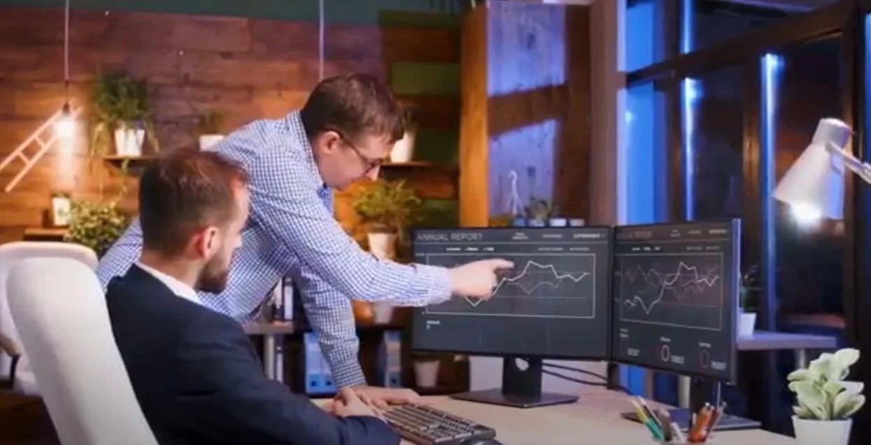 Two men working on a computer desk with dual monitors displaying financial graphs in a modern office.