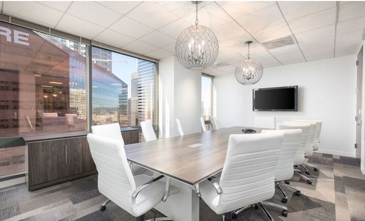 Modern conference room with white chairs around a wooden table, large windows with city views, wall-mounted TV, and two decorative pendant lights.