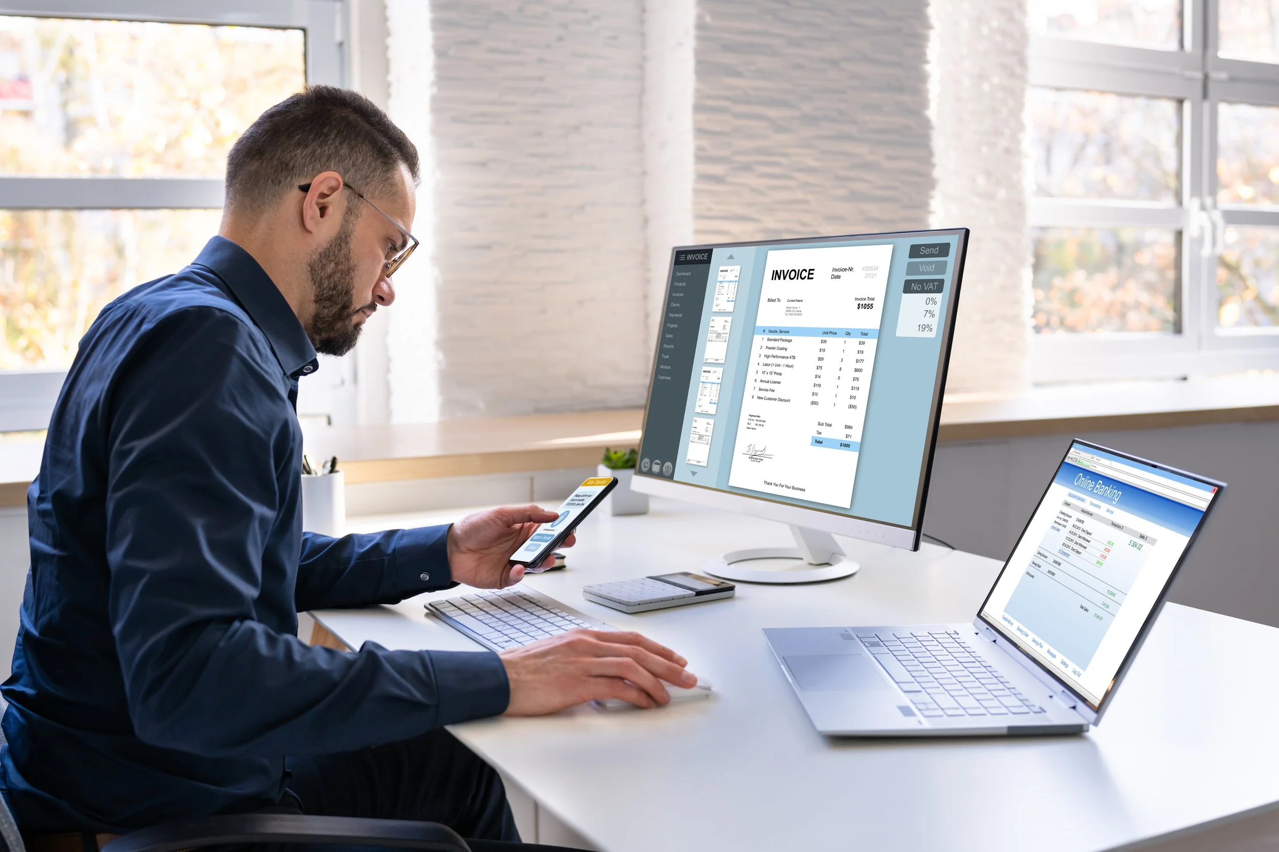 Man working at a desk with a computer monitor displaying an invoice, a laptop showing online banking, and a phone in hand.