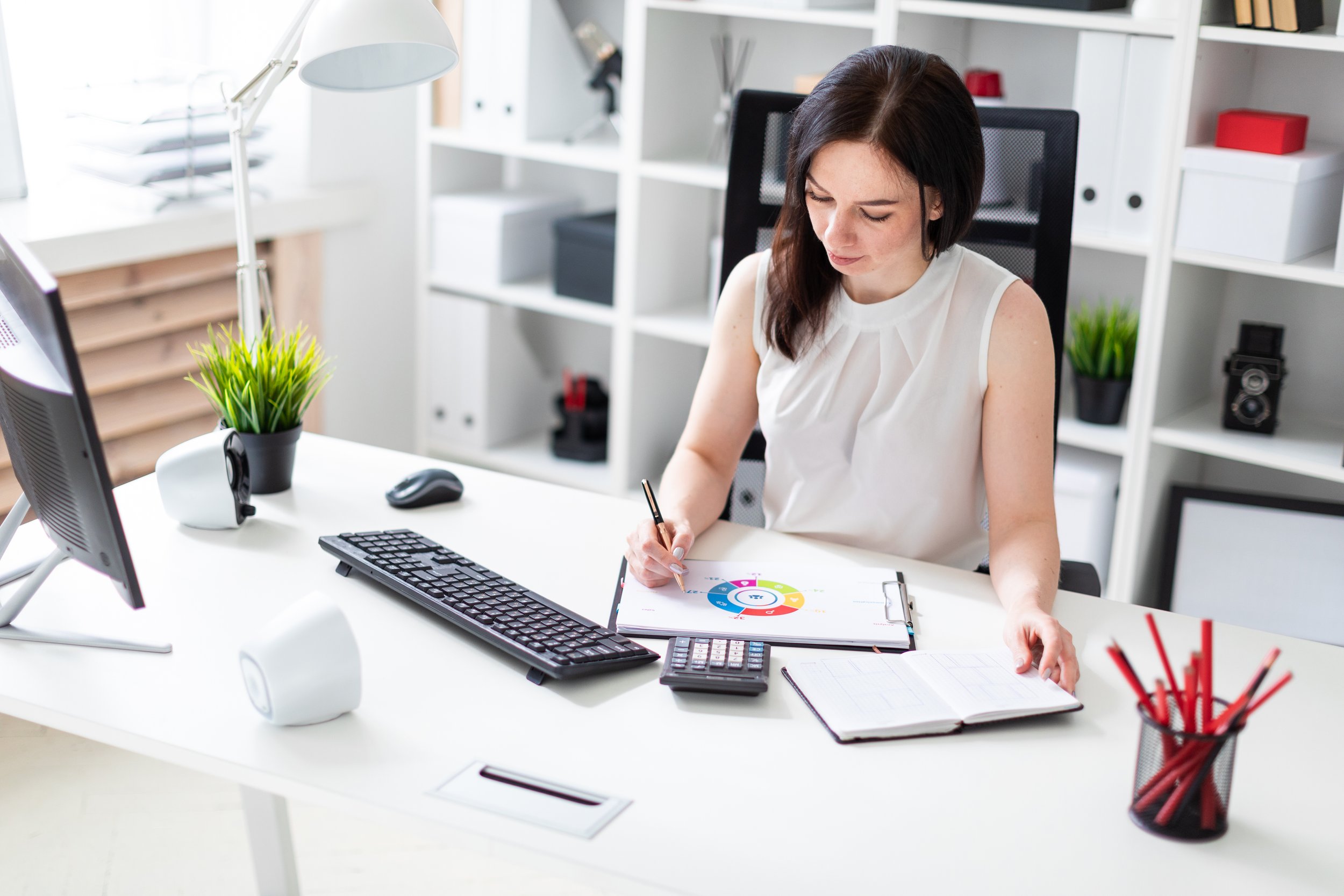 A woman sitting at a white desk, working with a pen on a colorful pie chart on a clipboard, surrounded by office supplies, with shelves and plants in the background.
