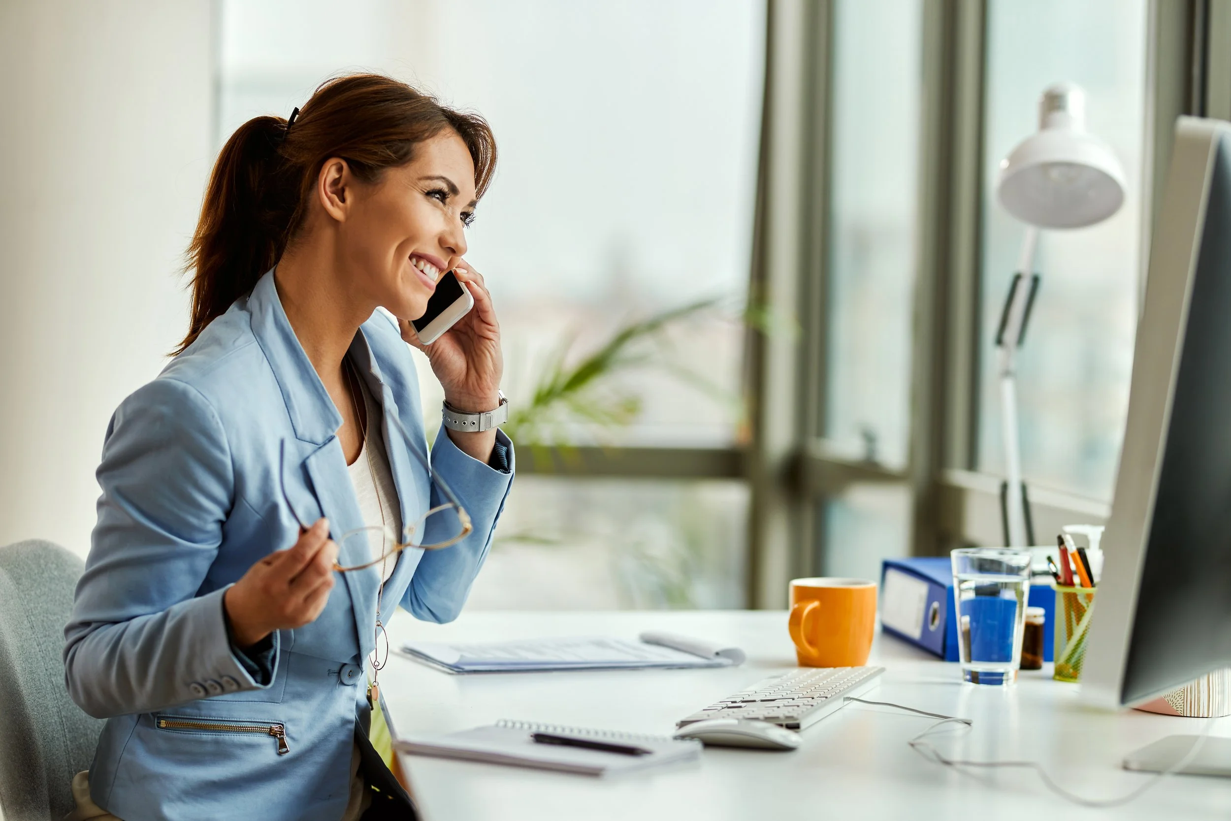 A woman with brown hair tied back, smiling while talking on a phone in a bright office. She is holding eyeglasses in one hand and standing near a desk with a keyboard, a notepad, a yellow coffee mug, a glass of water, and various office supplies. There is a large window and a lamp in the background.