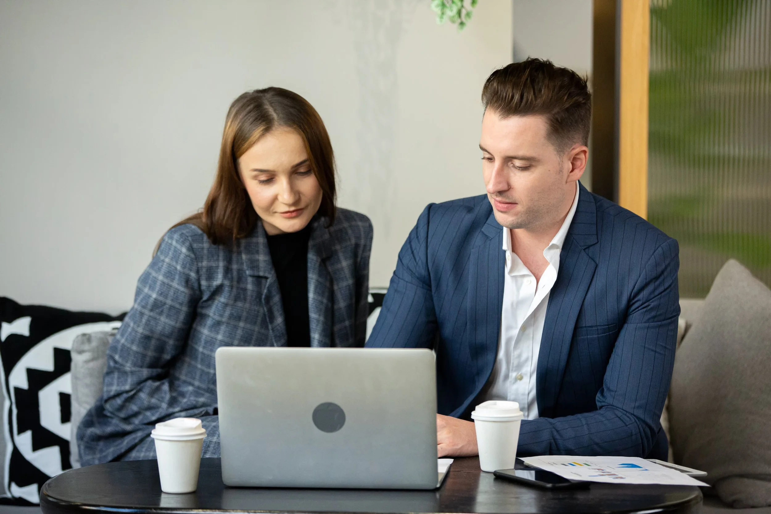 Two business professionals, a woman and a man, sitting closely at a table, looking at a laptop, with coffee cups and documents nearby, in a modern office setting.
