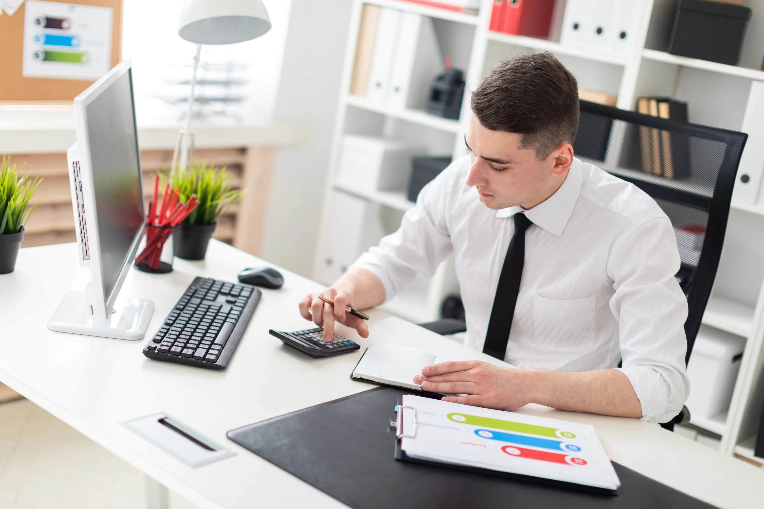 Young man in white shirt and black tie working at a modern office desk with computer, calculator, notepad, and colorful office supplies, in a bright office space with white furniture and shelves.