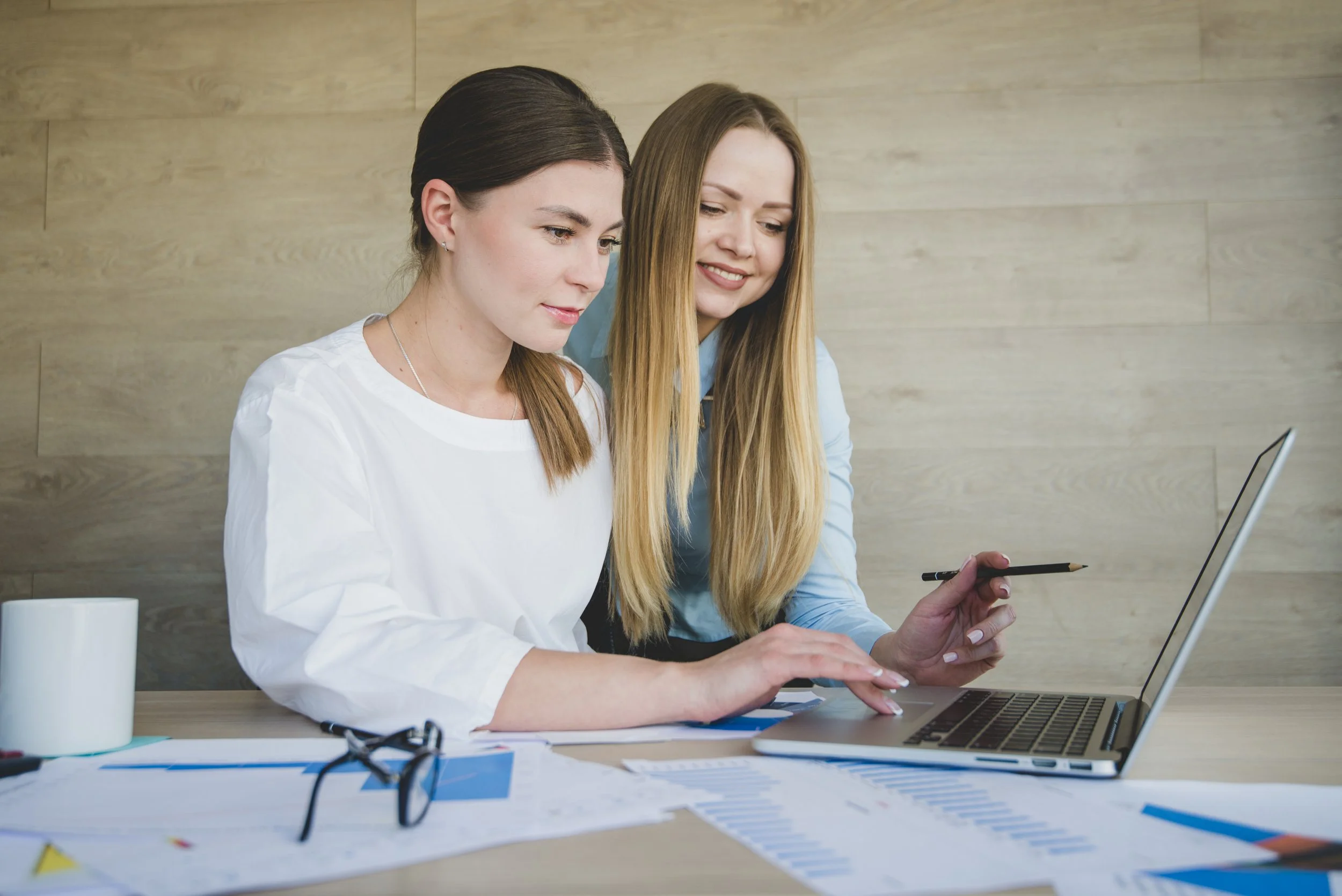 Two women working together at a desk with a laptop, papers, charts, and a coffee mug.