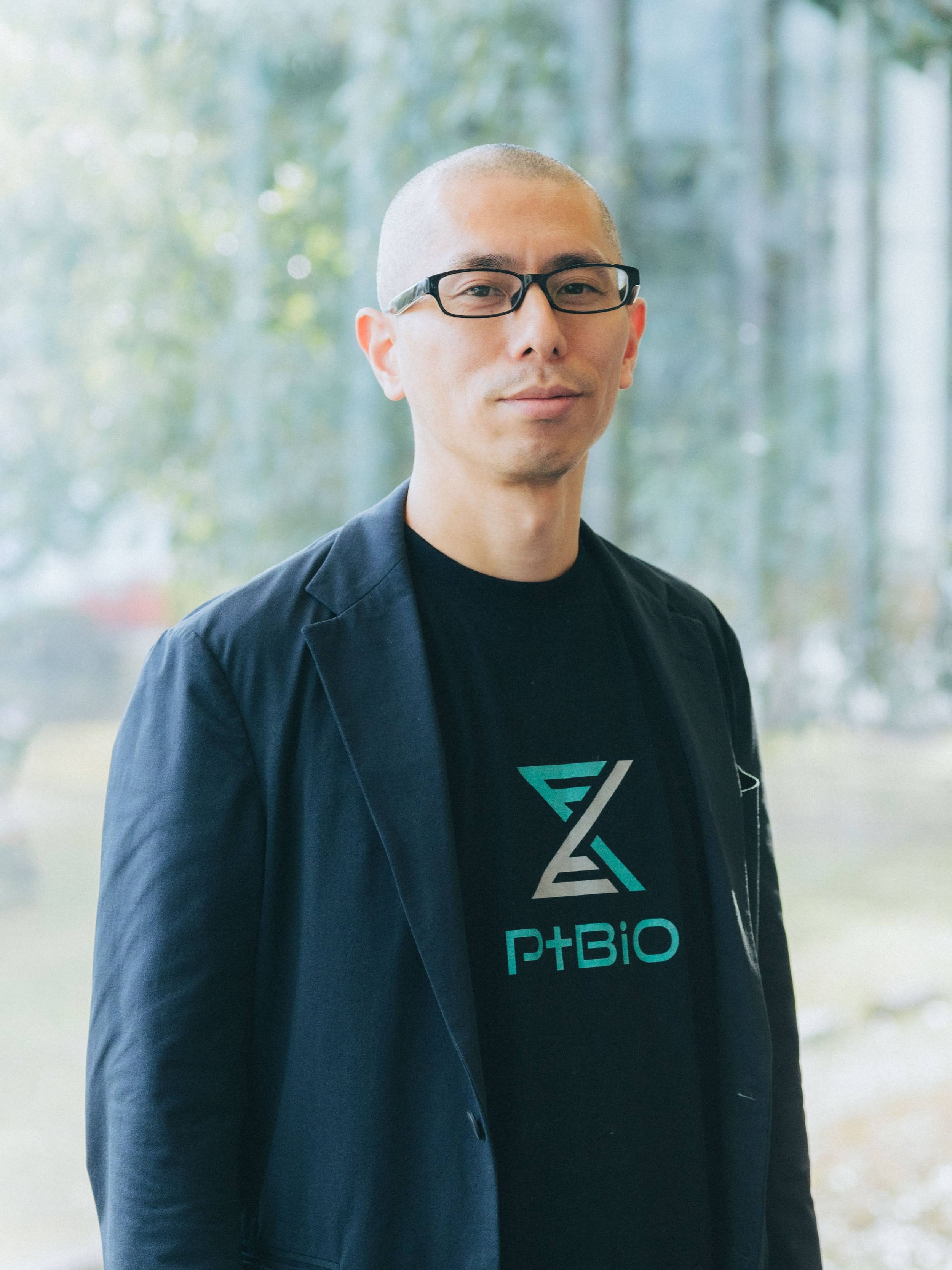 A man with glasses and a shaved head wearing a black T-shirt with a logo and text, standing outdoors with blurred trees in the background.