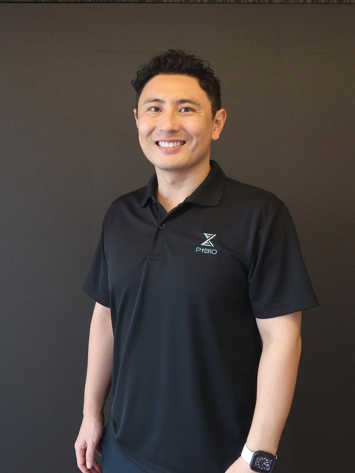 A smiling man with short dark curly hair, wearing a black polo shirt with a logo, standing against a plain dark background.