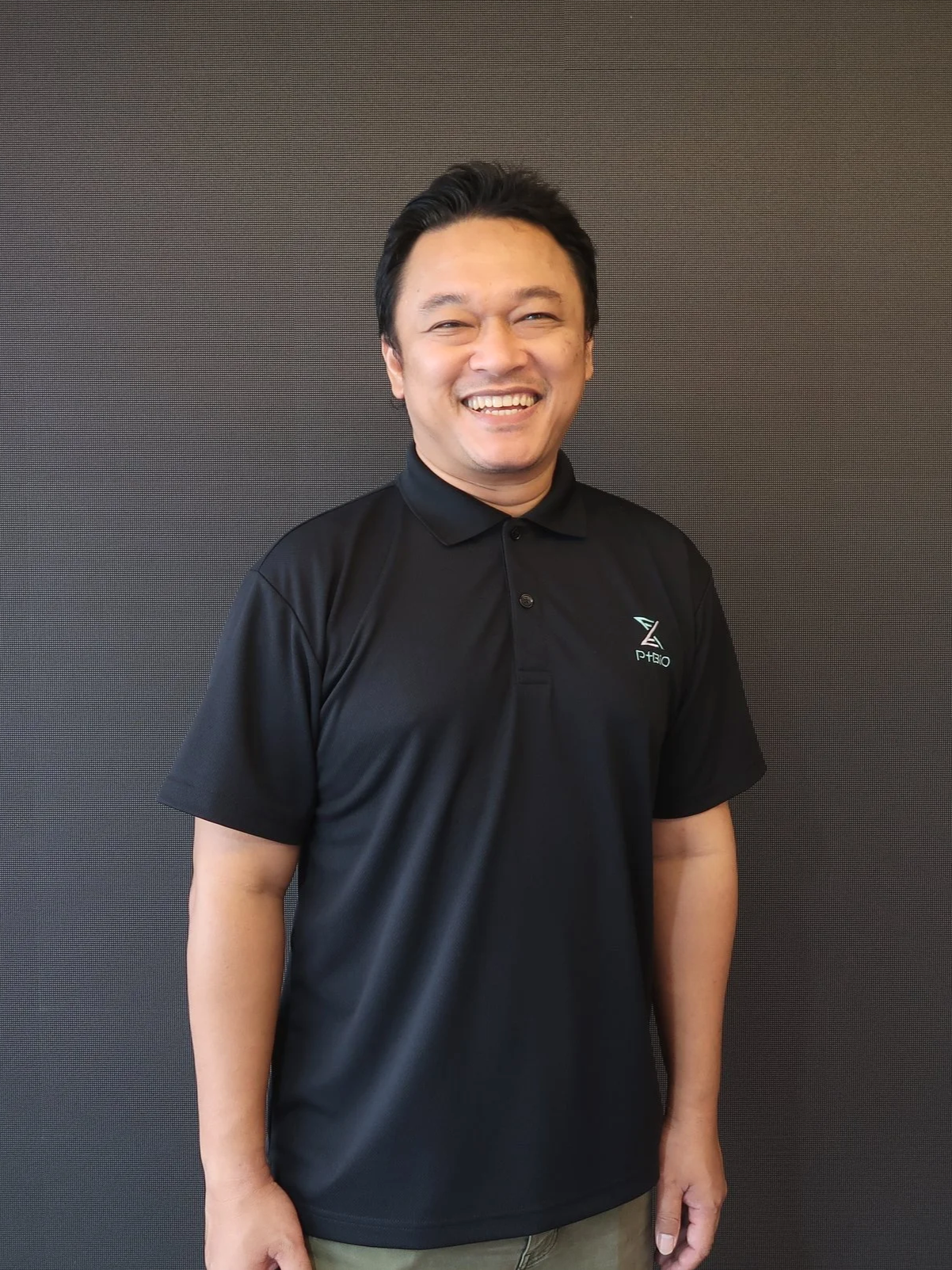 A smiling man with black hair wearing a black polo shirt standing against a dark background.