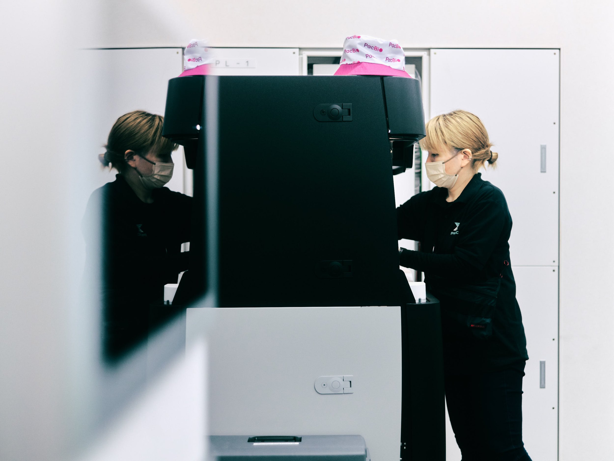 Two women wearing face masks working on a large 3D printer or similar machine in a lab or tech environment.