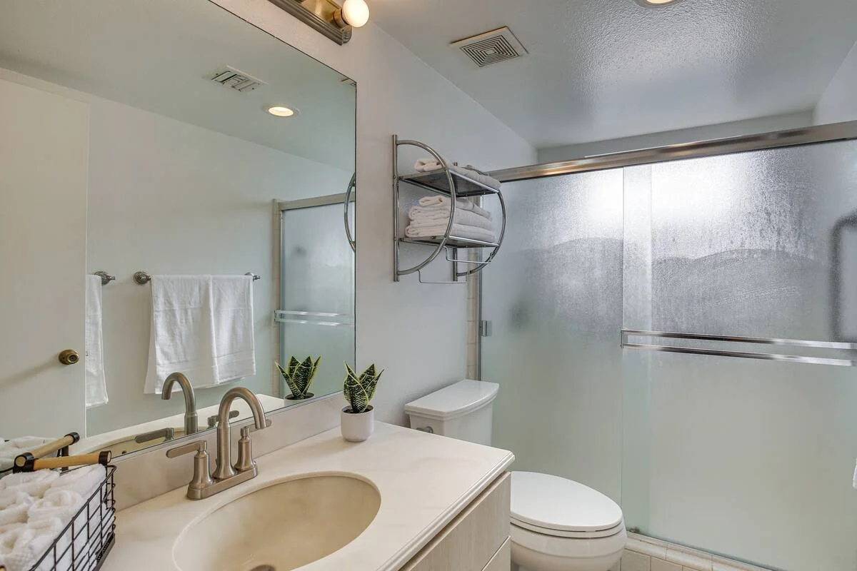 Clean bathroom with white walls, a large mirror, a cream-colored vanity with two plants, a toilet, a towel rack with white towels, and a glass shower door.