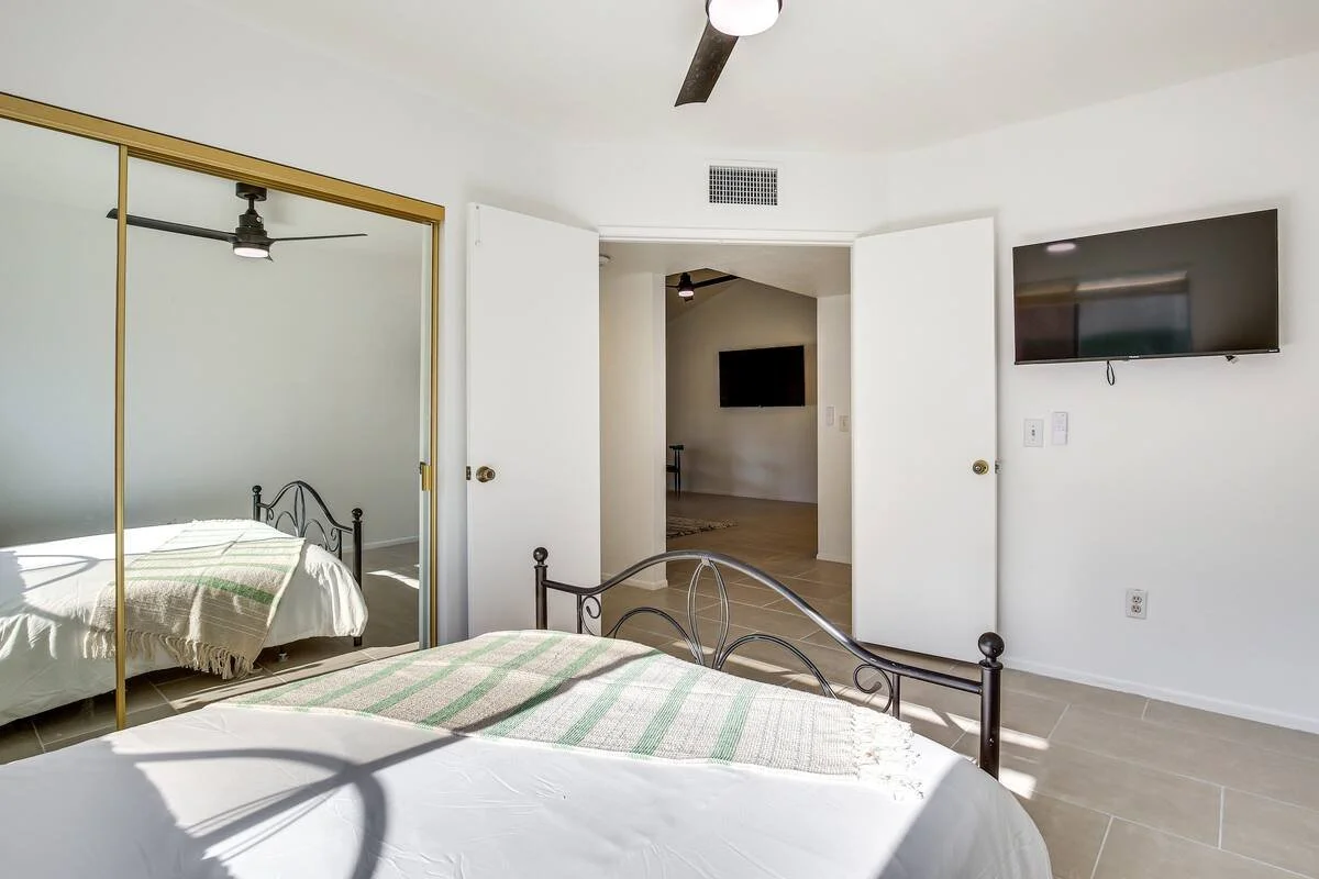 Bedroom with a bed, mirrored closet doors, a TV mounted on the wall, and a door leading to another room, with sunlight streaming in.