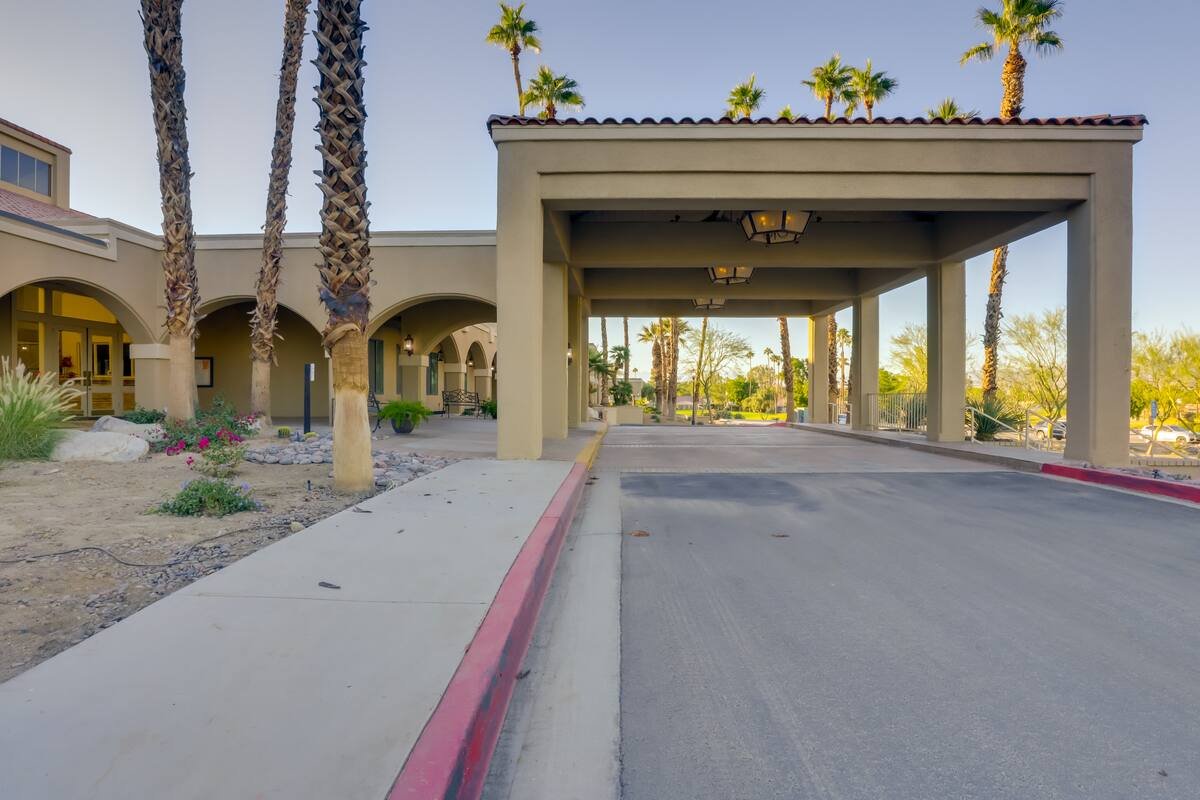 Covered entrance of a building with palm trees and a sidewalk on the left, and a road with a red curb on the right, in a sunny setting.
