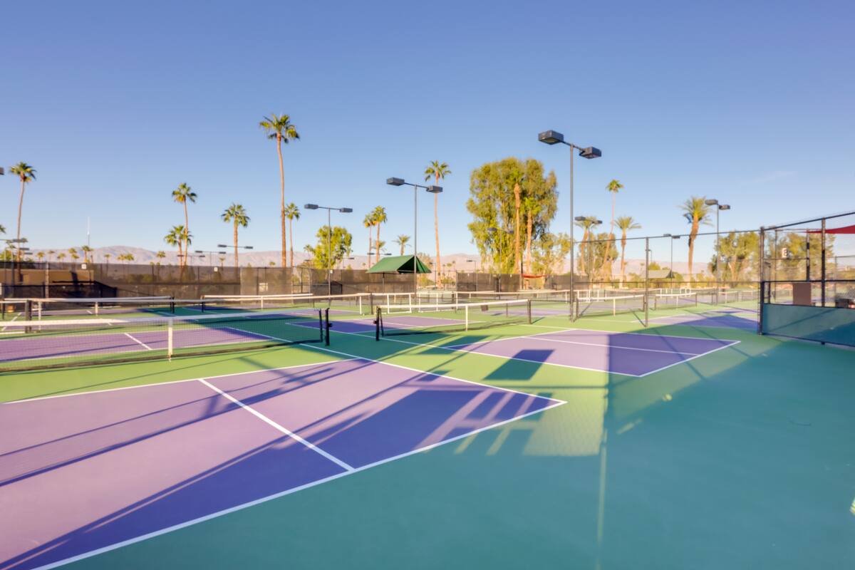 Multiple tennis courts with purple and green surfaces, surrounded by a chain-link fence, with palm trees and clear blue sky in the background.