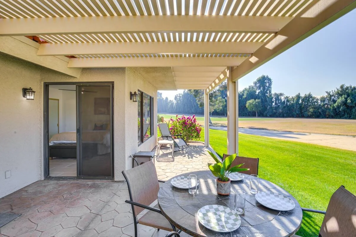 Covered patio with outdoor dining table set with plates and glasses, planted with a green plant in the center, overlooking a grassy yard with trees in the distance.