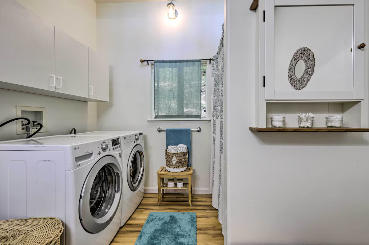 Laundry room with white LG washer and dryer, wooden floor, small window with curtain, and shelves with decorative items.
