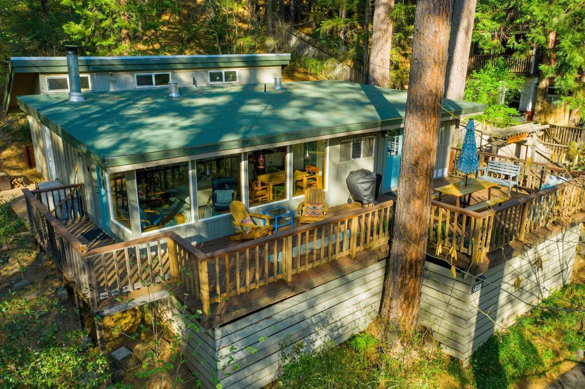 A raised house with a green roof and large windows, surrounded by tall trees and a wooden deck with outdoor furniture and an umbrella.