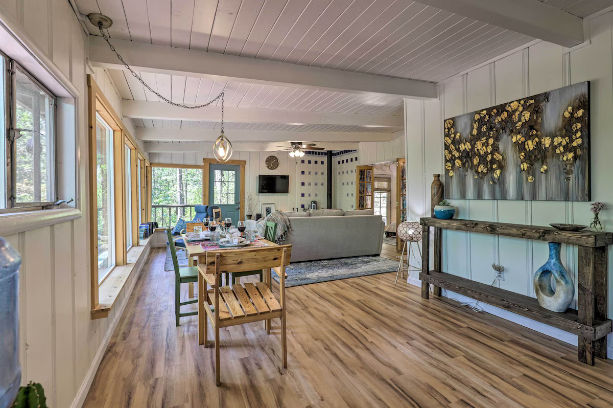 Spacious living and dining area with wood flooring, light-colored walls, large windows, a dining table set, various furniture, and decorative wall art.