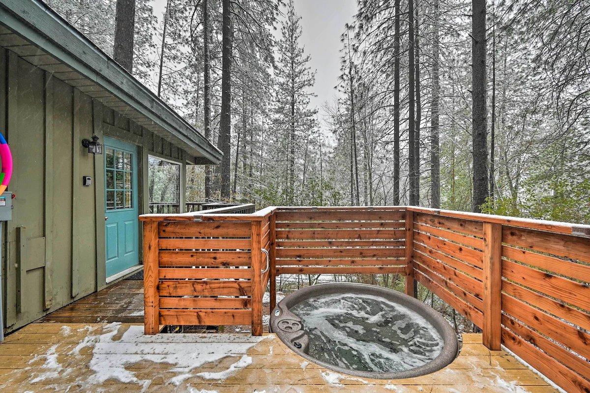 Outdoor hot tub on a wooden deck surrounded by a wooden fence, with snow and ice, and tall trees in the background.