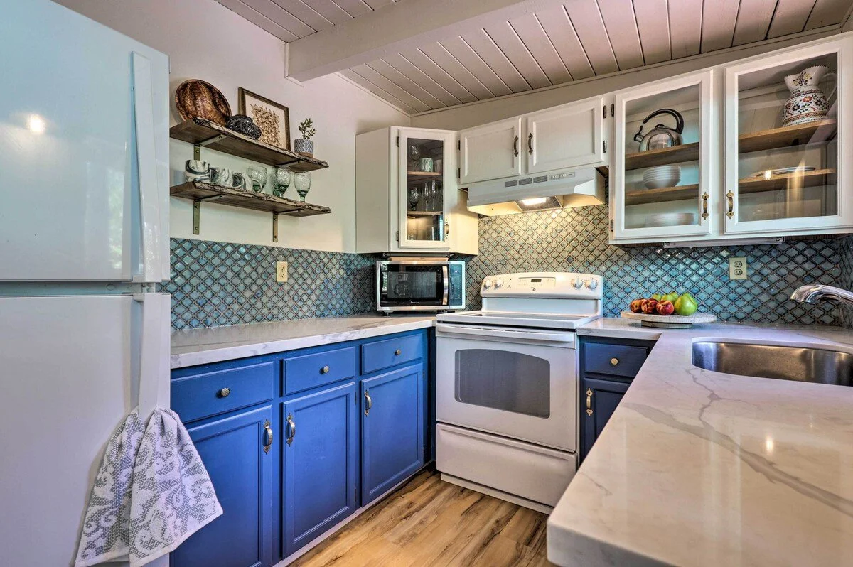 Kitchen with white upper cabinets, dark blue lower cabinets, a white stove, a microwave, open shelves with dishes and decorative items, a bowl of fruit on the counter, and a stainless steel sink.