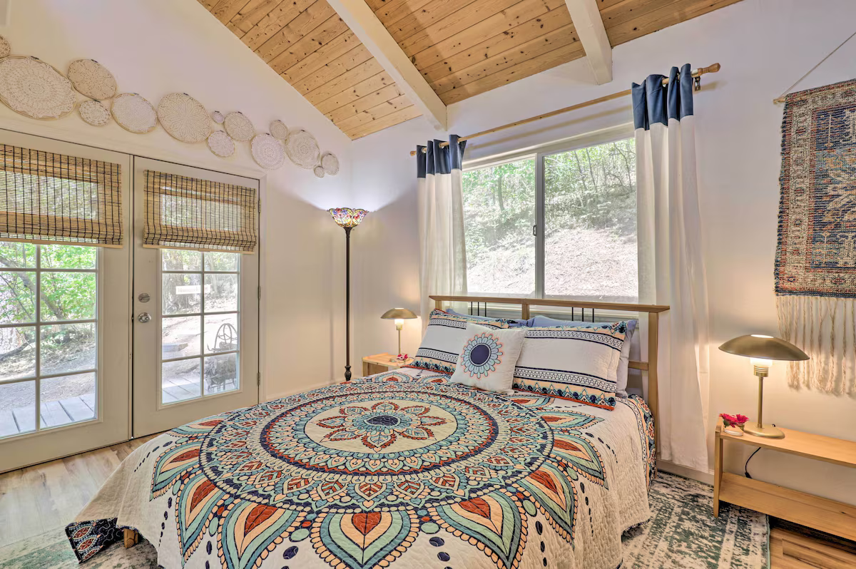 A cozy bedroom with a wooden ceiling, decorated with round woven wall hangings, featuring a bed with a colorful mandala-patterned quilt, surrounded by lamps, curtains, and a view of greenery outside through large windows.