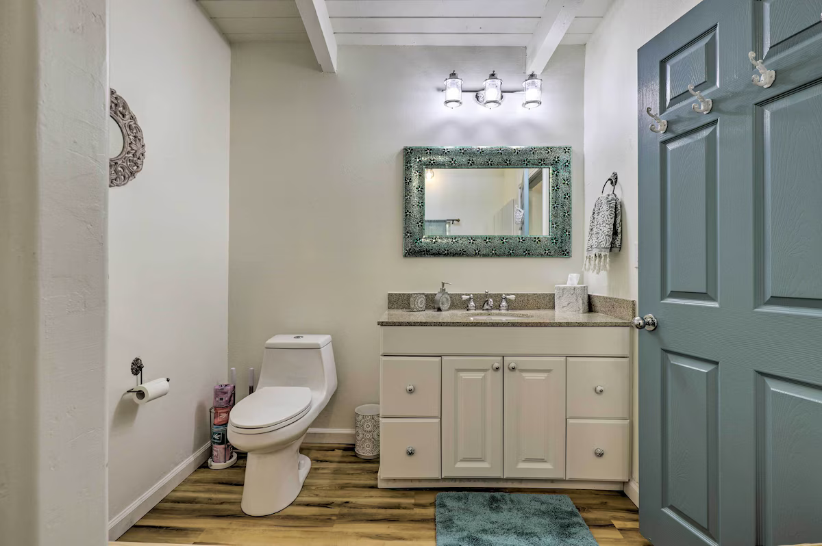 Bathroom with a white toilet, a cream vanity with a granite countertop, a decorative mirror, a towel hanging on the wall, and a blue door with hooks. The room has wood flooring and a ceiling with white beams.