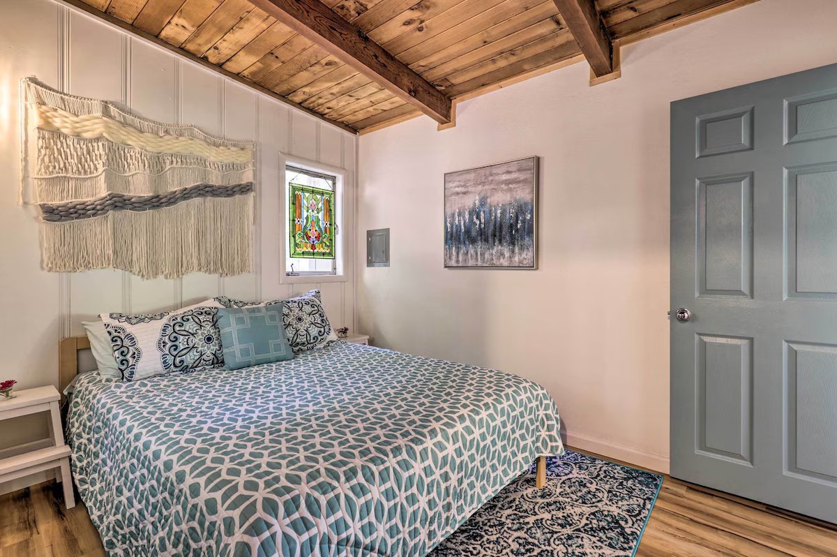 Bedroom with a bed covered in a patterned bedspread, decorative pillows, a woven wall hanging, a stained glass window, a painting, and a wooden ceiling.