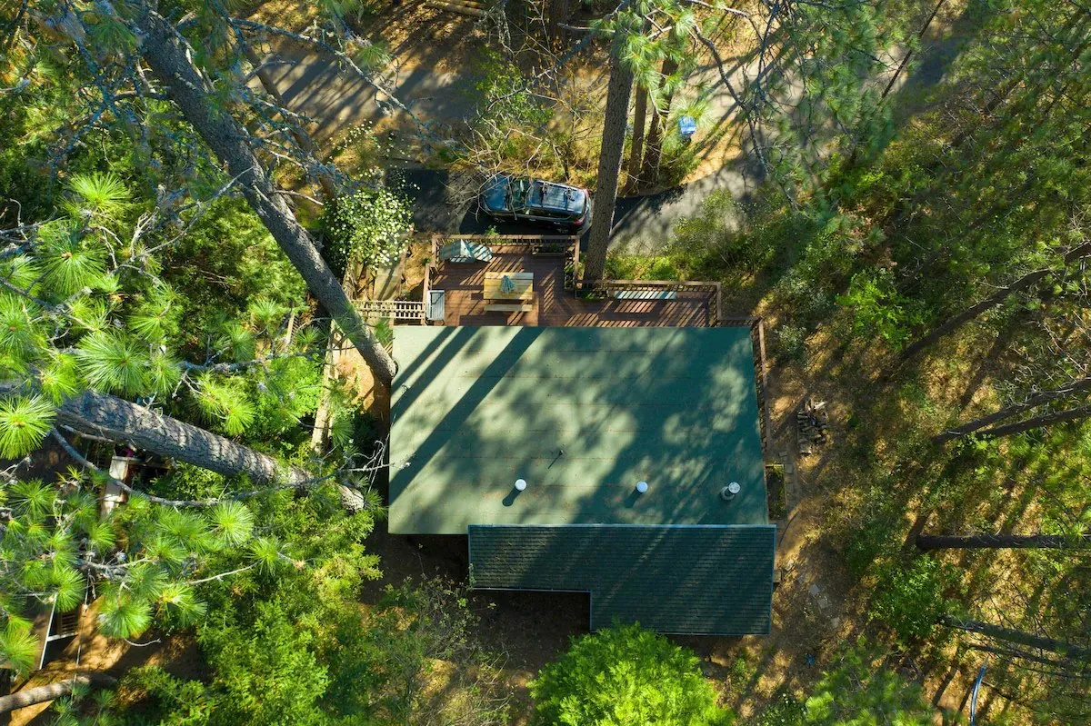 Aerial view of a house surrounded by tall trees with a green roof and a wooden deck area with outdoor furniture.