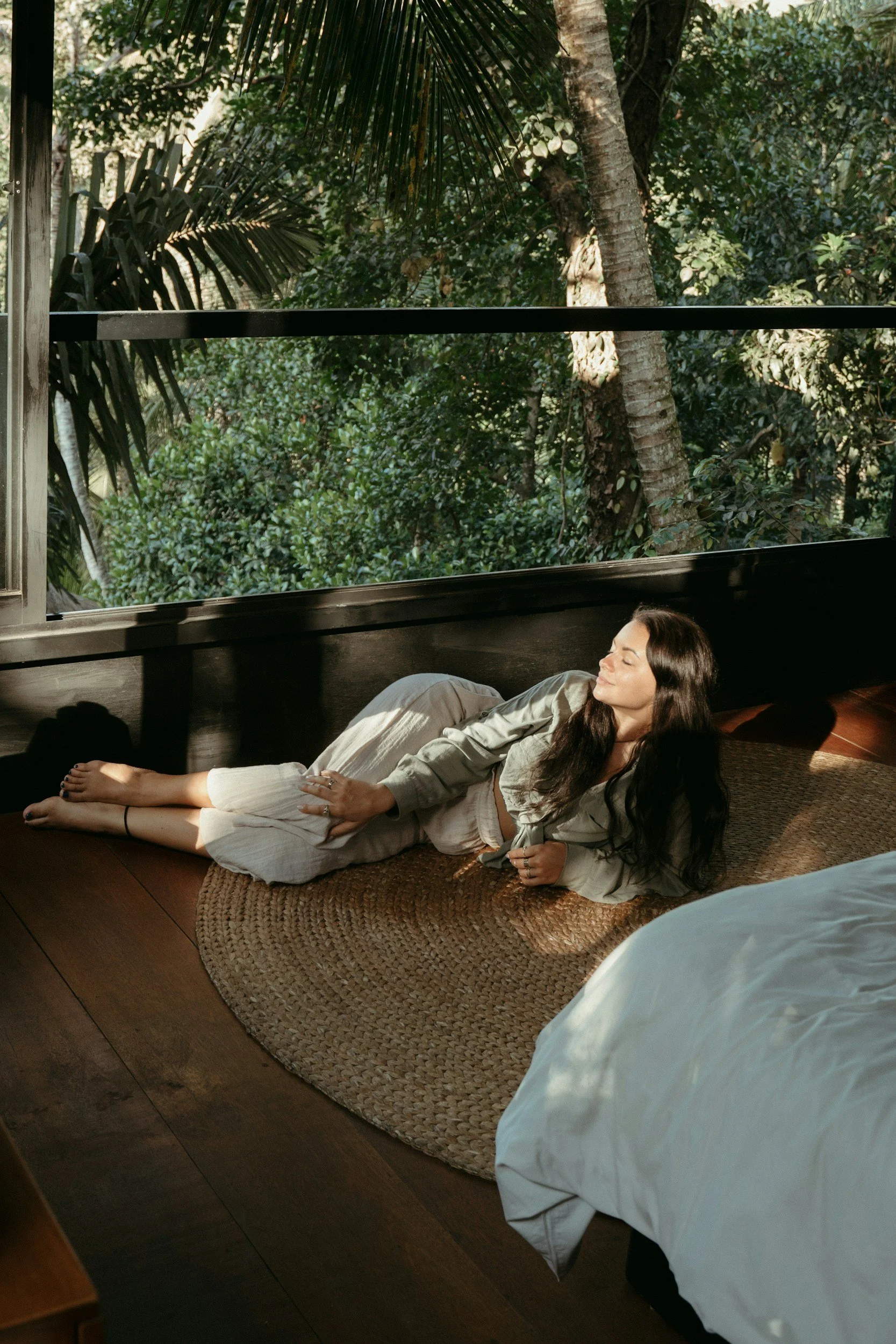 A woman lying on a woven rug on a wooden floor, basking in sunlight coming through large open windows. She has long dark hair, is wearing a light-colored relaxed outfit, and appears to be enjoying the peaceful outdoor view of greenery and trees.