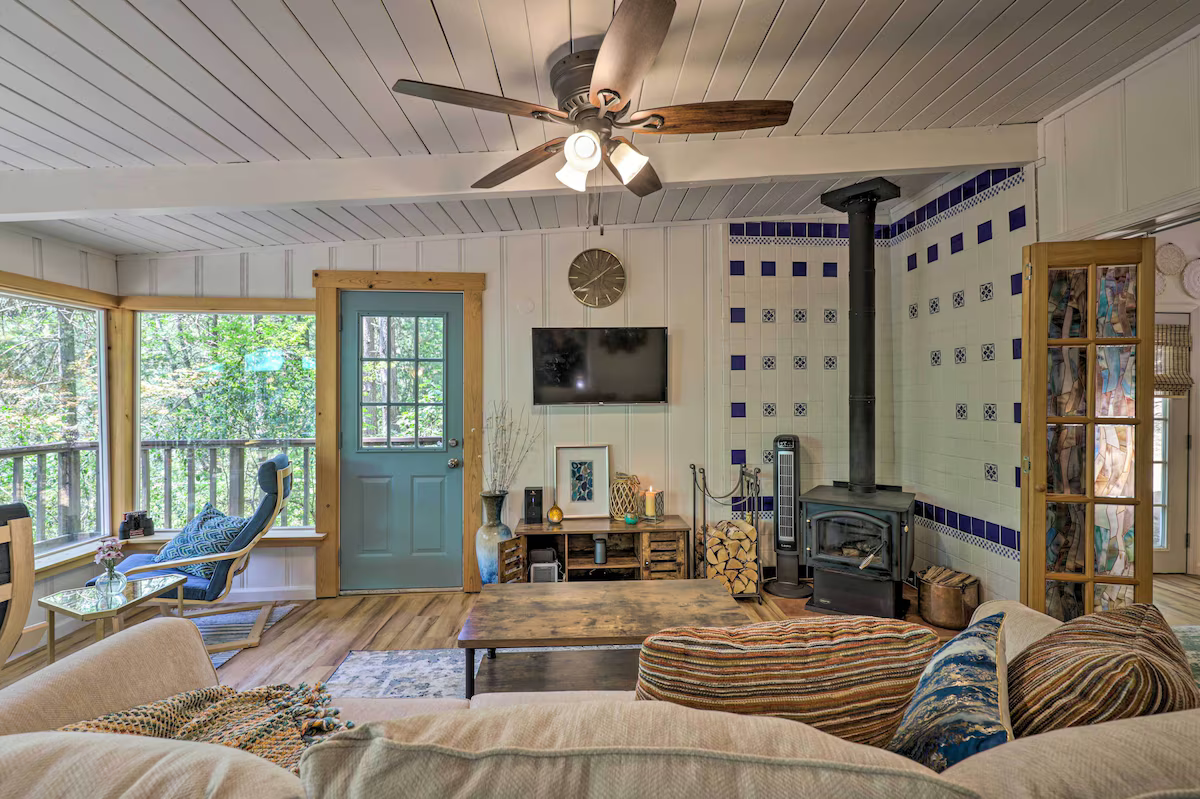 Living room with a ceiling fan, wood stove, wall-mounted TV, large windows, and a cozy seating area.