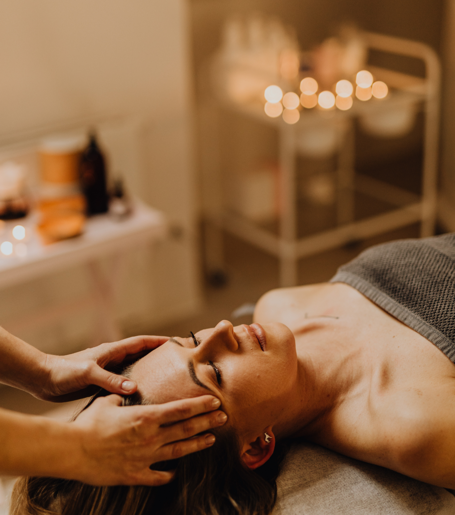 A woman receives a facial massage in a spa setting, lying on a massage table with her eyes closed, while a therapist's hands gently press her temples.