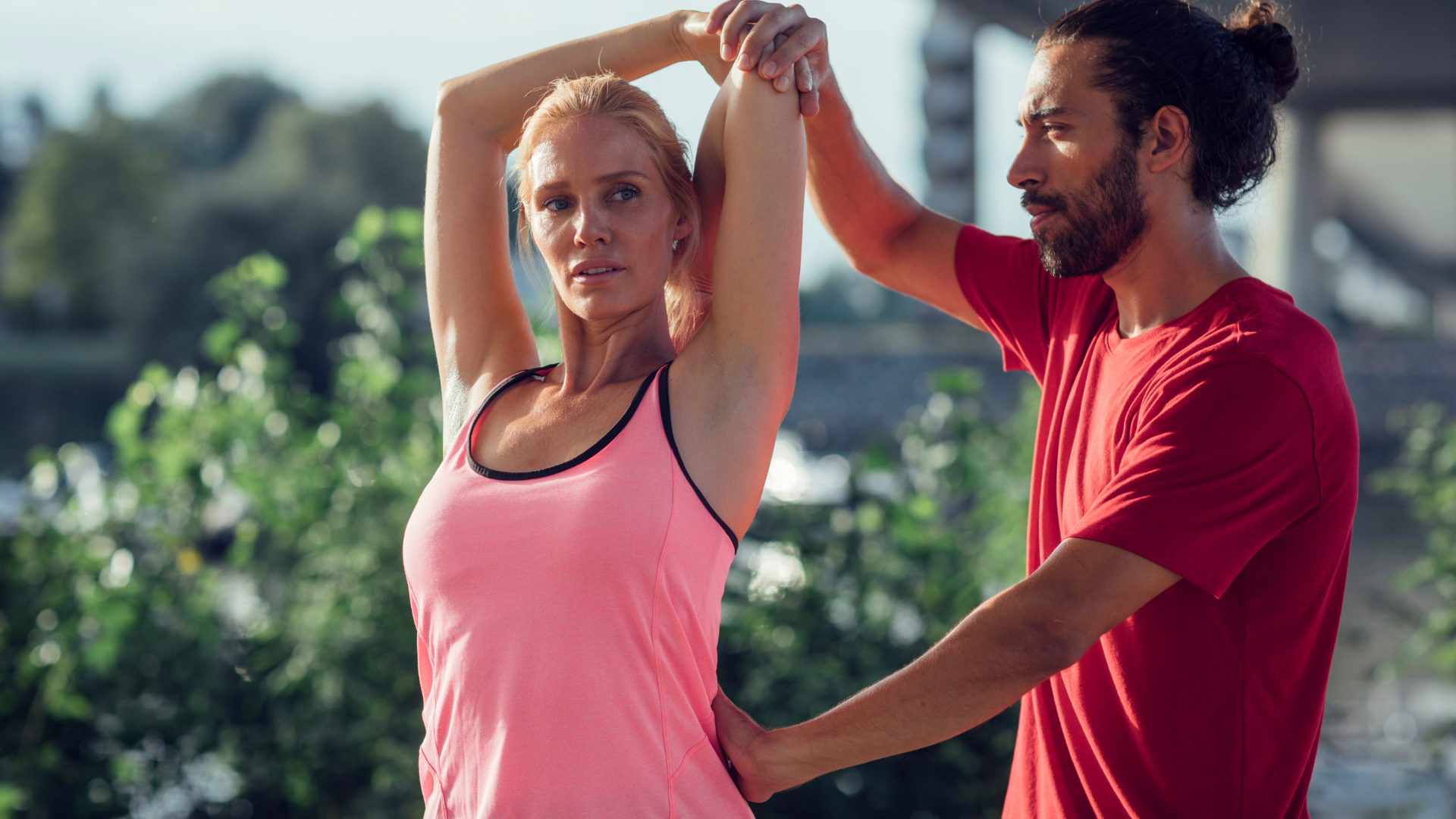 A woman stretching outdoors with a man assisting her, both are dressed in athletic clothing.