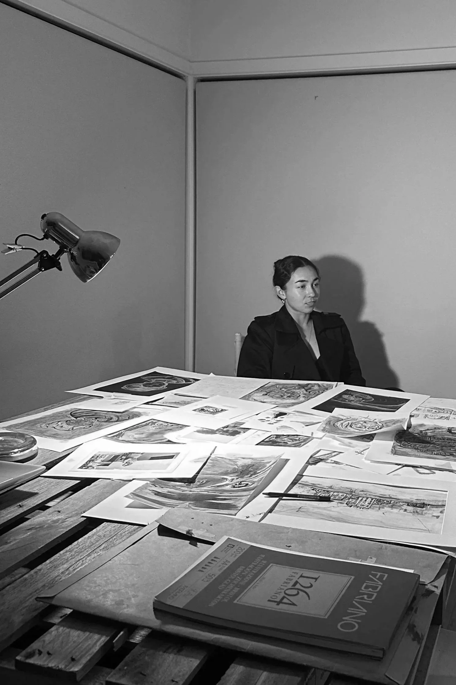 A woman sitting at a table covered with papers and artwork in a black and white photo. There is a desk lamp on the left side and a closed book in the foreground.