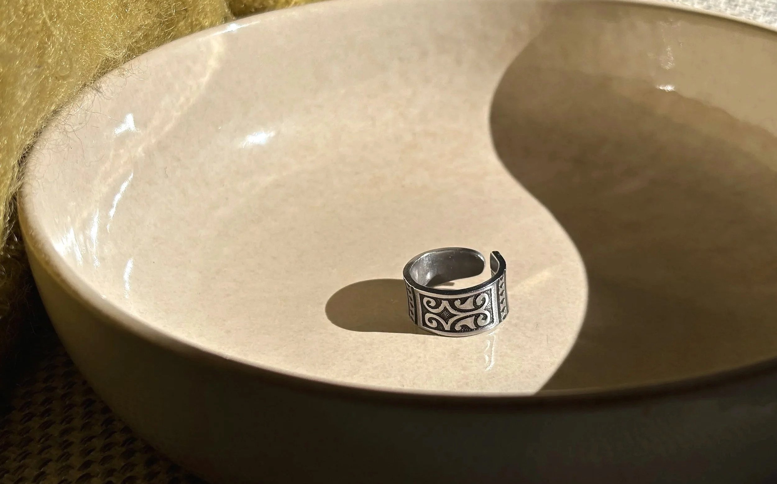 A silver ring with intricate black swirling patterns rests inside a cream-colored, shallow ceramic dish, casting a shadow on the dish's inner surface.