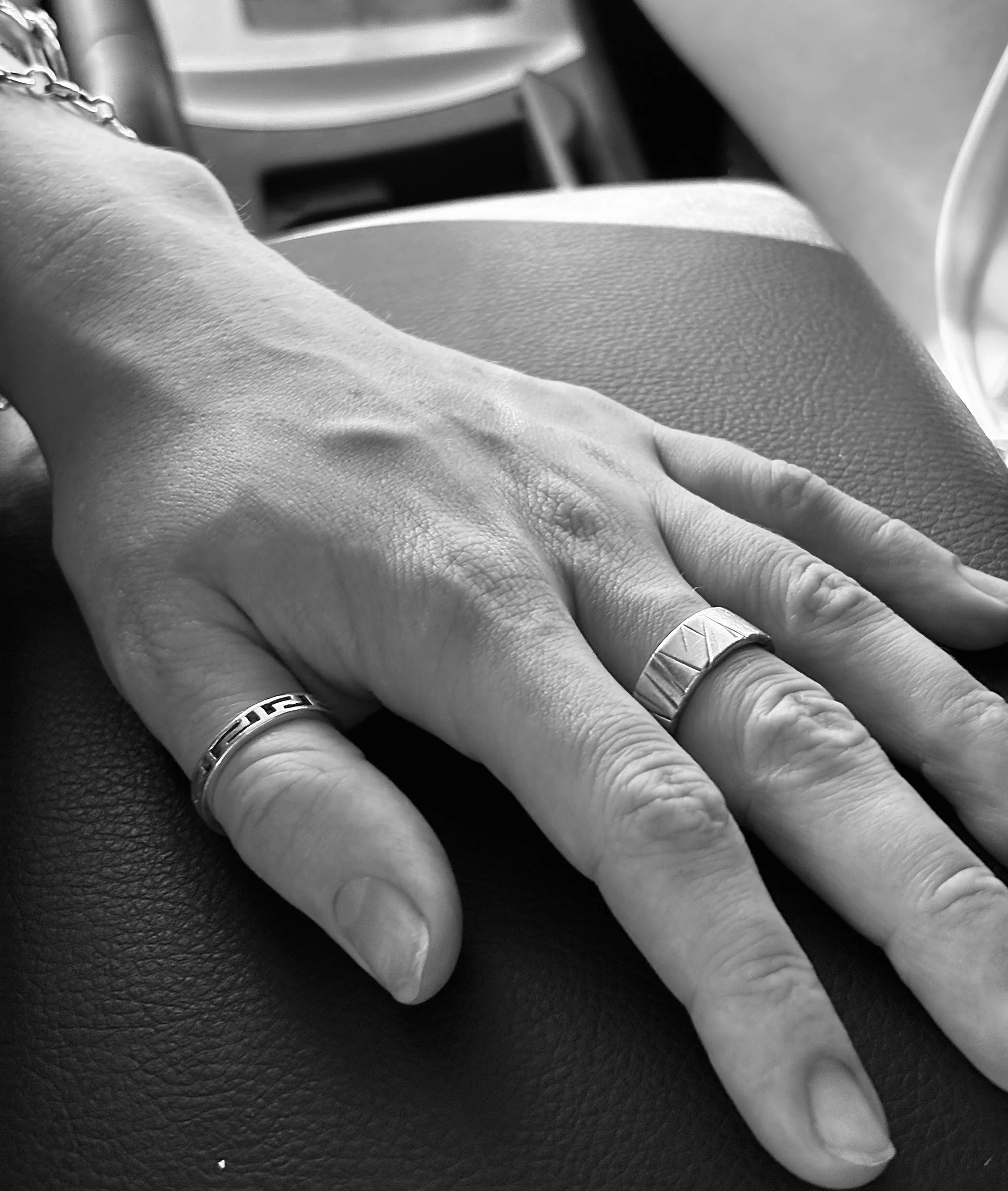 A close-up of a person's hand resting on a textured surface, wearing two rings, one on the middle finger and one on the ring finger, in black and white.