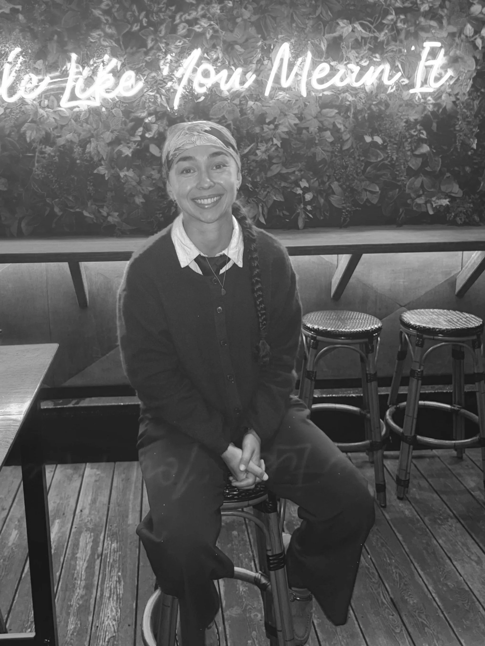 A woman with a braid wearing a headscarf, a jacket, and a collared shirt, smiling and sitting on a stool in a café or restaurant with a neon sign and leafy wall in the background.