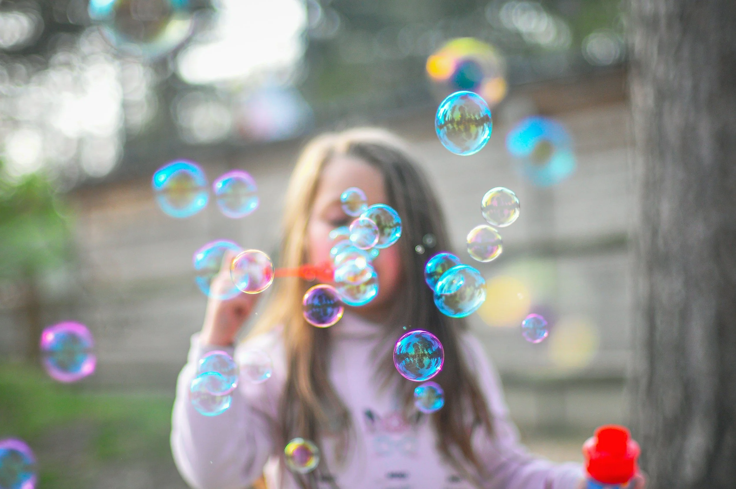 A young girl blowing bubbles outdoors, with many colorful bubbles floating in the air and a blurred background including a fence, trees, and a tree trunk.