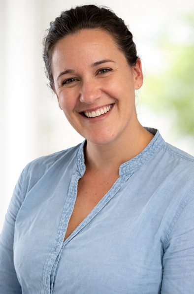 A woman with dark brown hair smiling, wearing a light blue blouse, with a bright background of blurred greenery.