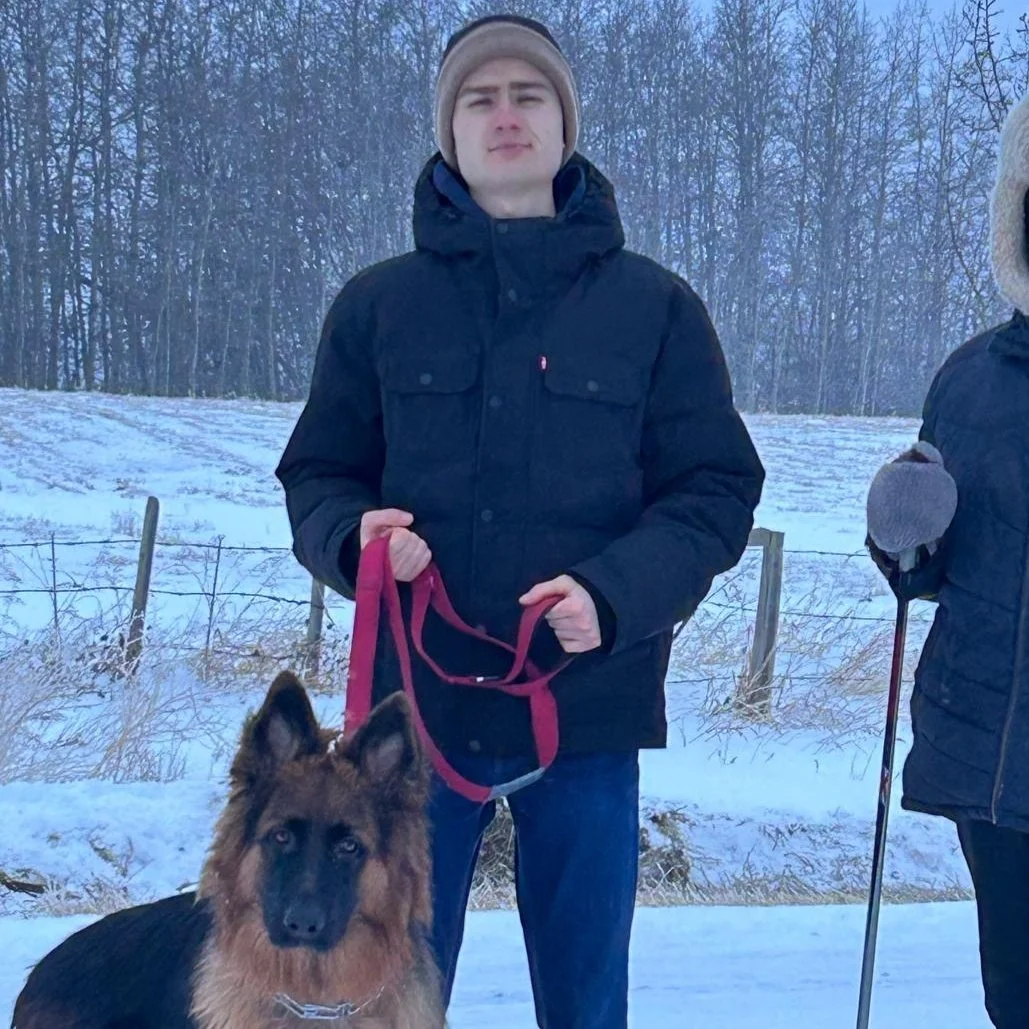 A young man in a black winter coat and beige beanie stands outdoors in a snowy field, holding a red leash attached to a German Shepherd dog. The background shows a snow-covered landscape with trees.