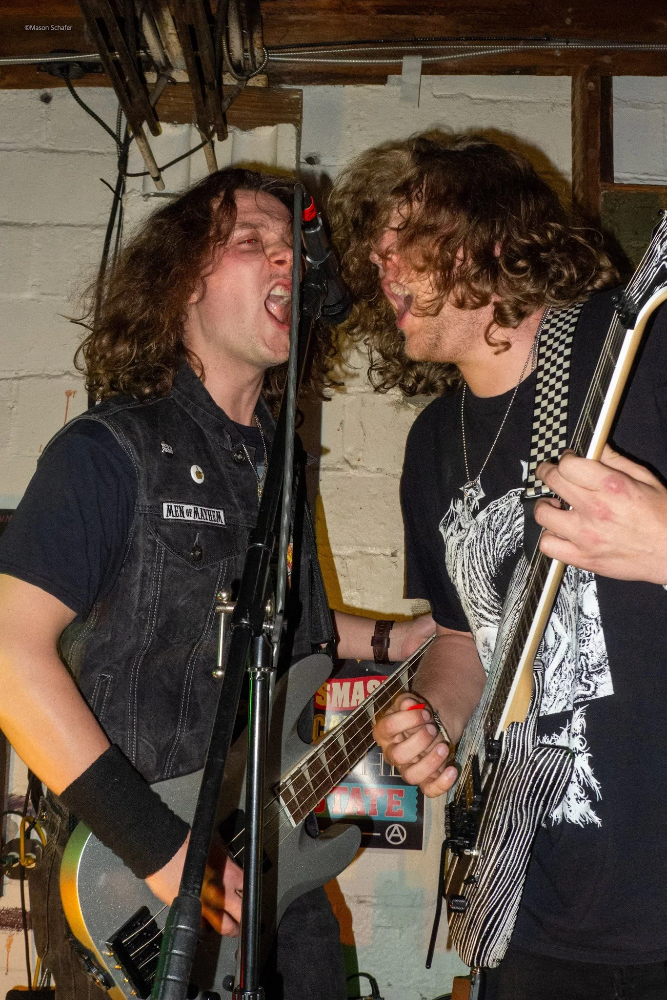 Two men passionately singing and playing electric guitars in a band practice space; both with long curly hair, one wearing a vest with a patch reading 'Men of Mayhem' and the other in a band t-shirt, close to each other with mouths open, shouting or 