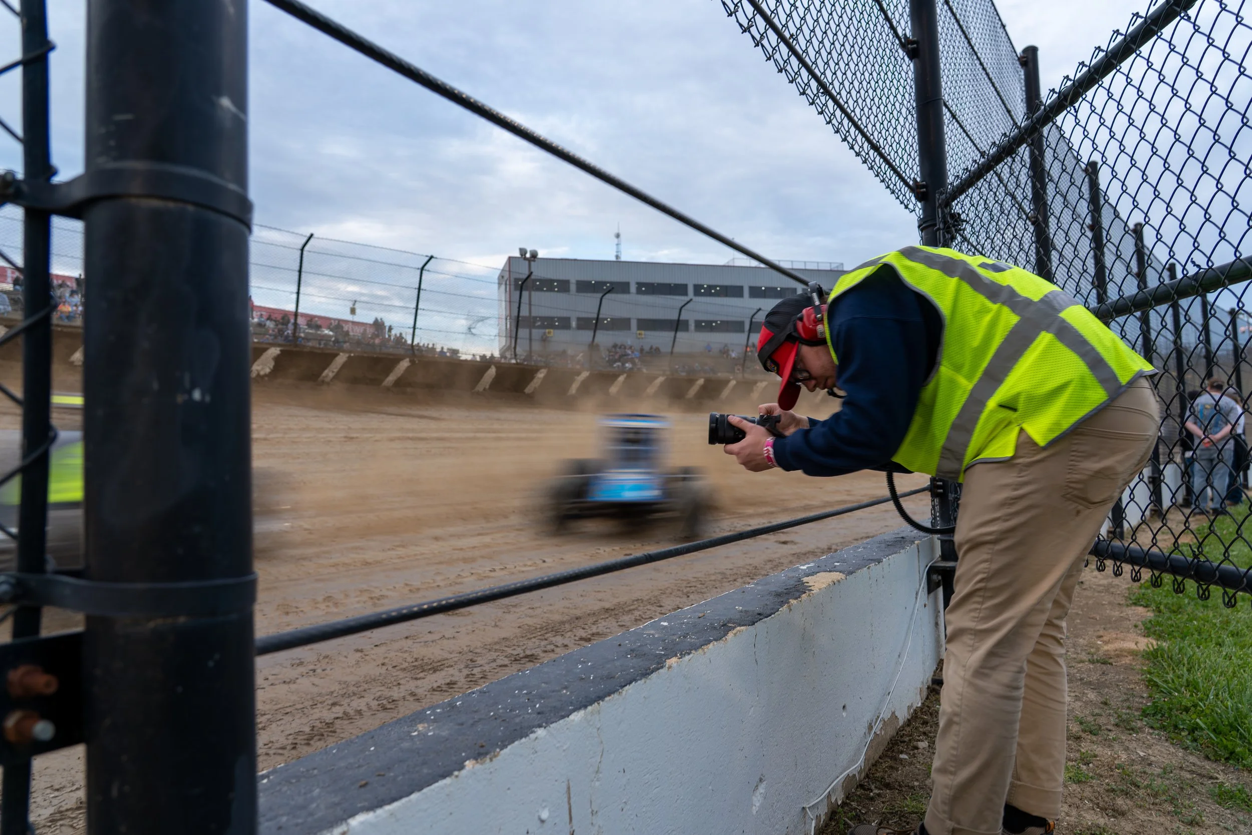 Photographer kneeling near a racetrack, wearing a yellow safety vest and red cap, taking a picture of a race car on the dirt track, with a chain-link fence and a crowd in the background.