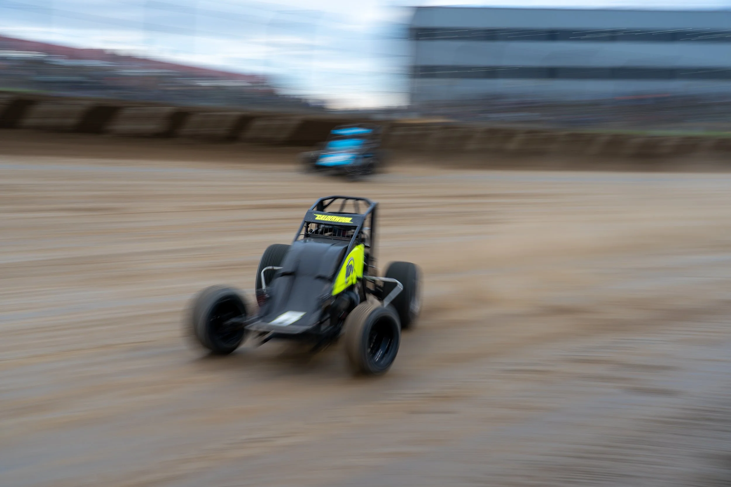 A black and yellow racing go-kart speeding on a dirt track during a race with a blurred background of spectators and buildings.