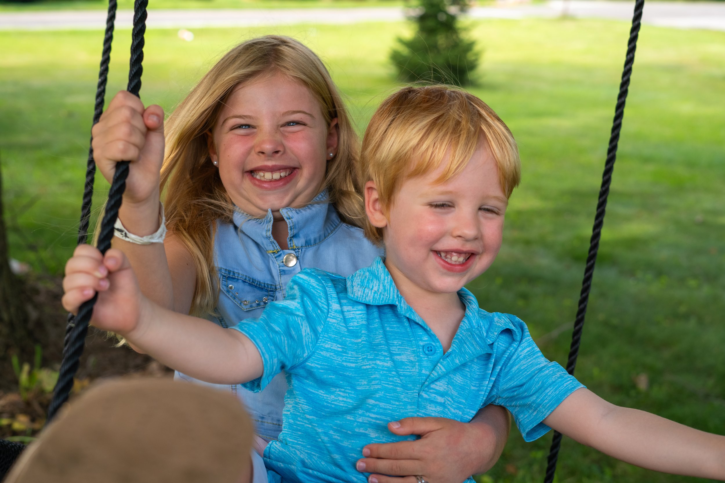A young girl and boy smiling while sitting on a swing set outdoors in a park with green grass and trees in the background.