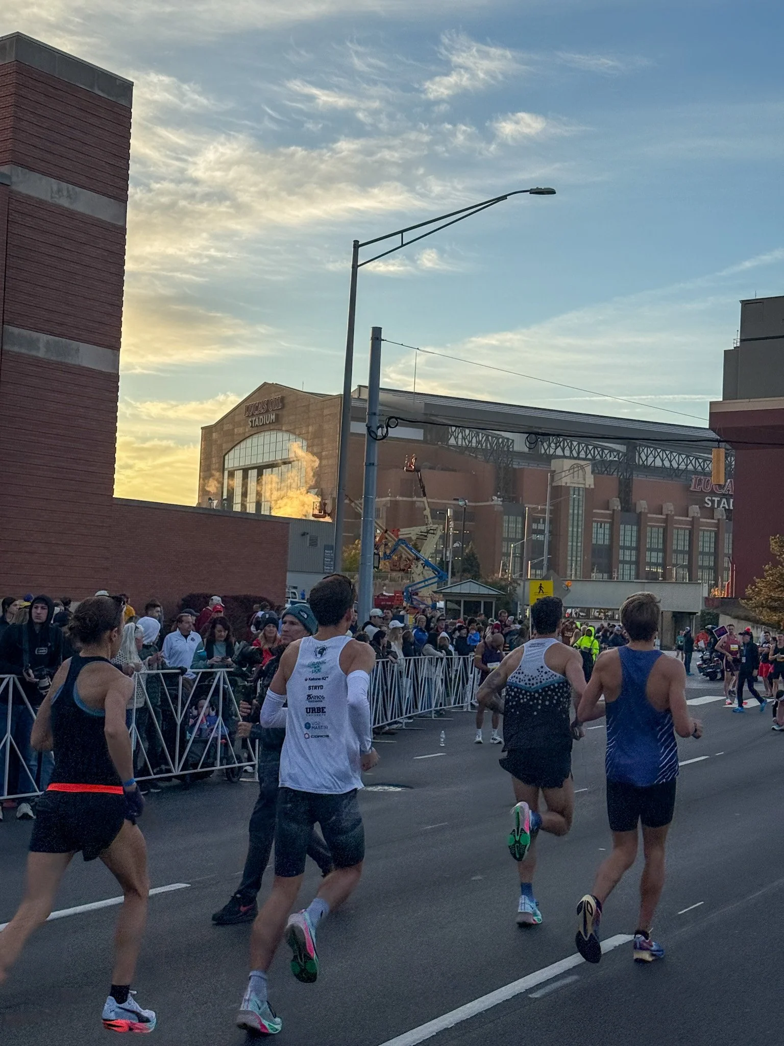 Marathon runners competing on city street near Lucas Oil Stadium during sunset with spectators behind barriers.