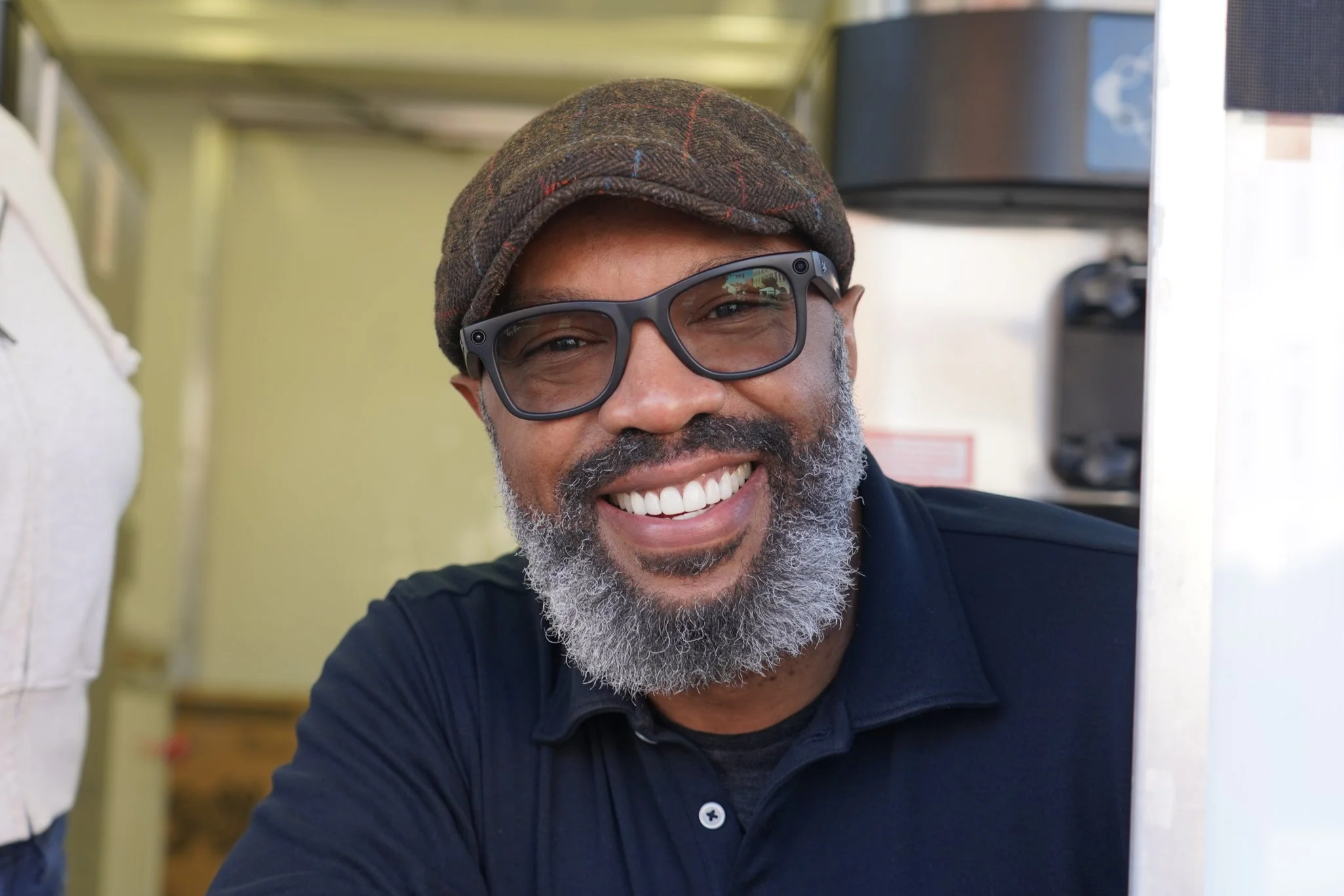Man with gray beard and glasses smiling, wearing a brown cap, in a setting with kitchen appliances.