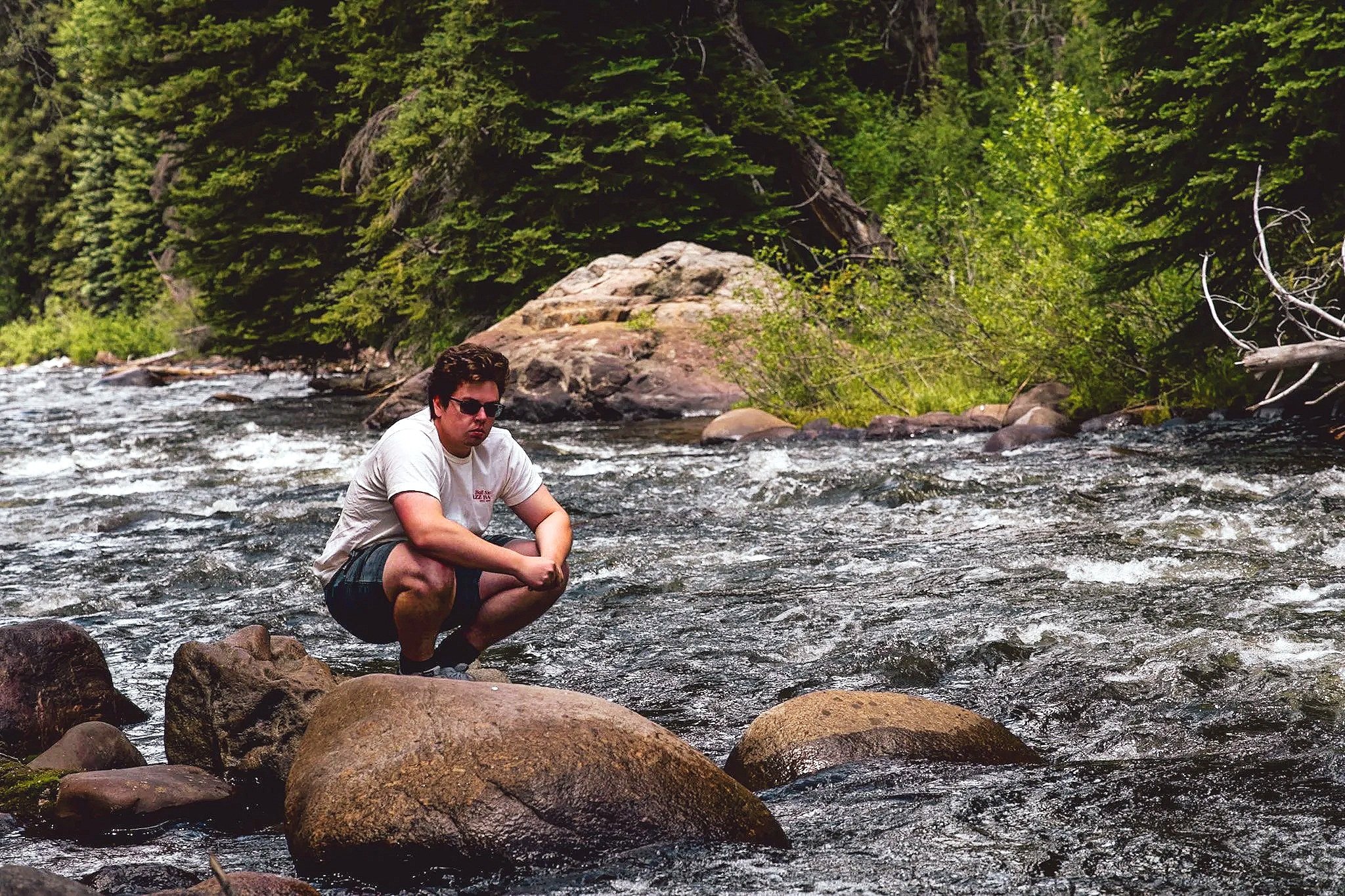 A man squatting on rocks in a flowing river with lush green trees and bushes in the background.