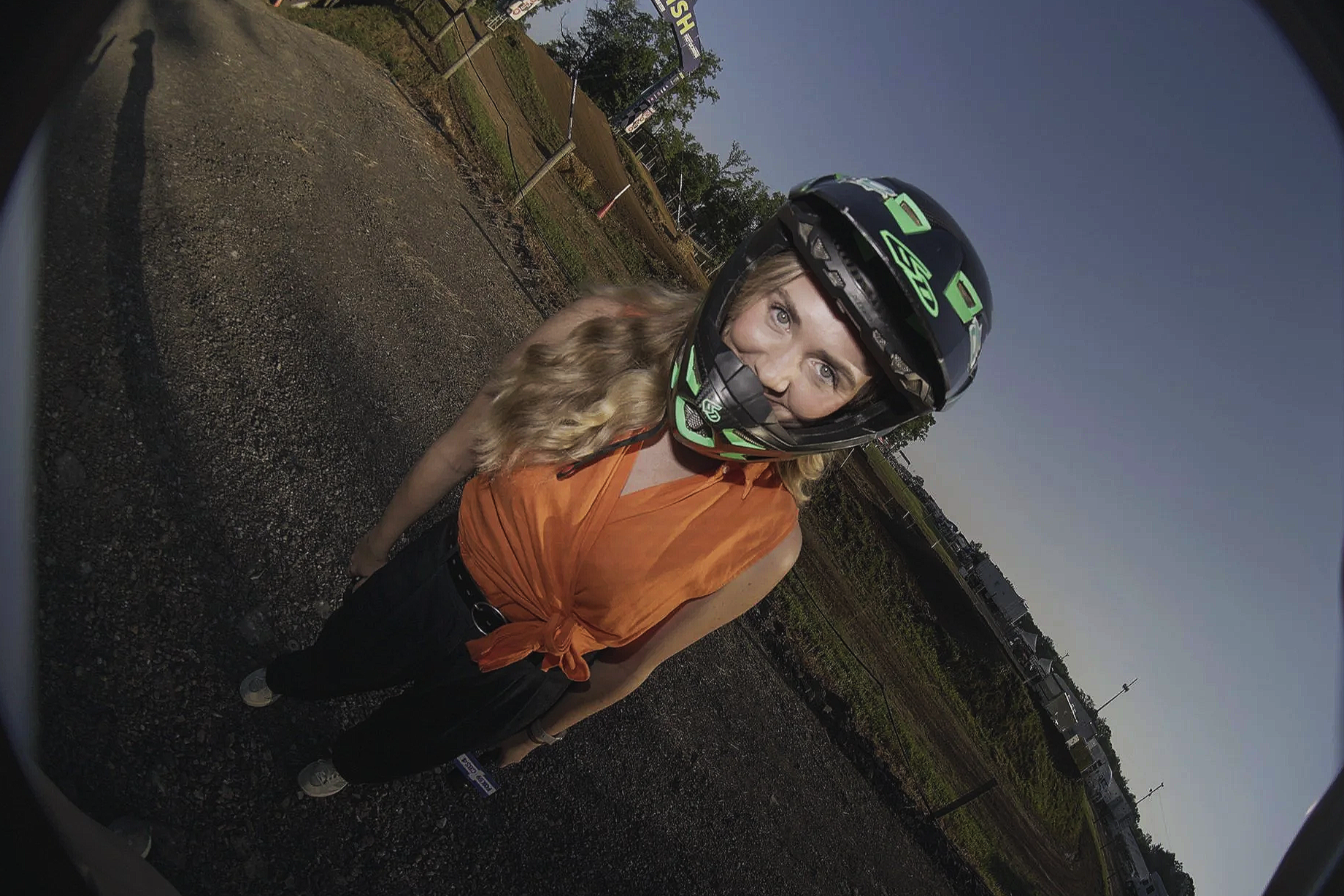 A woman with long blonde hair wearing a black and green motorcycle helmet, an orange sleeveless top tied at the waist, black pants, and white sneakers. She is standing outdoors on a gravel area with fields and power lines in the background during lat