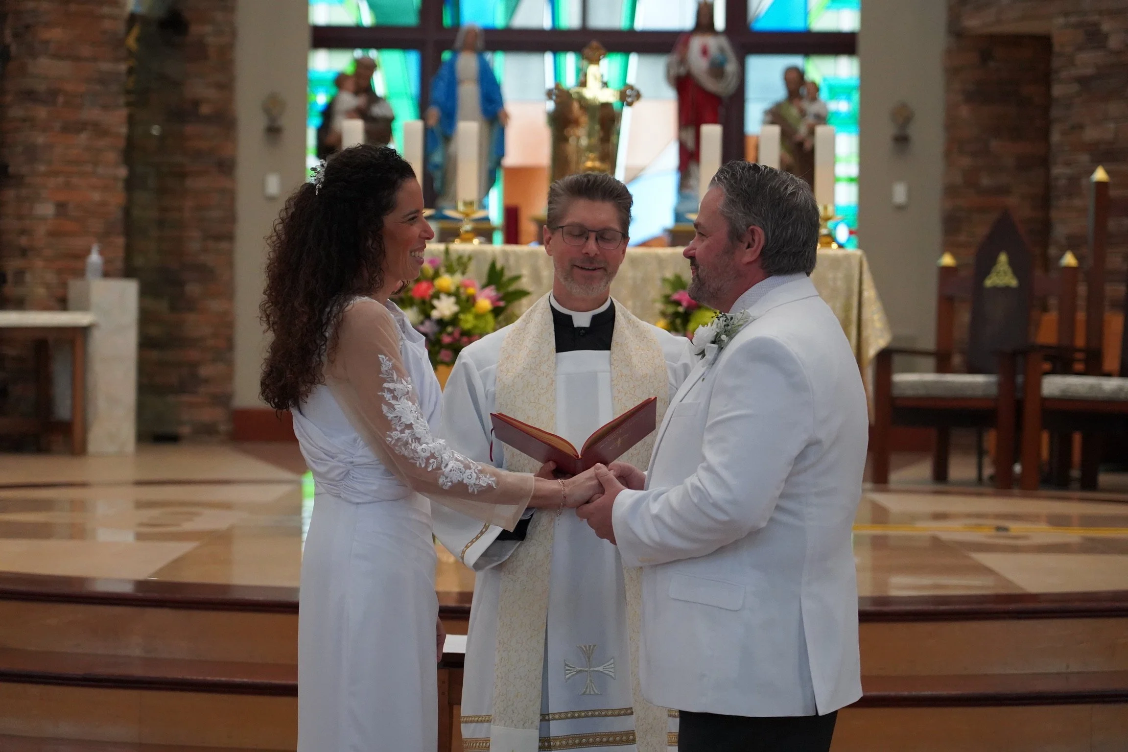 A bride and groom exchange vows during their wedding ceremony in a church, with a priest standing between them and holding a book.